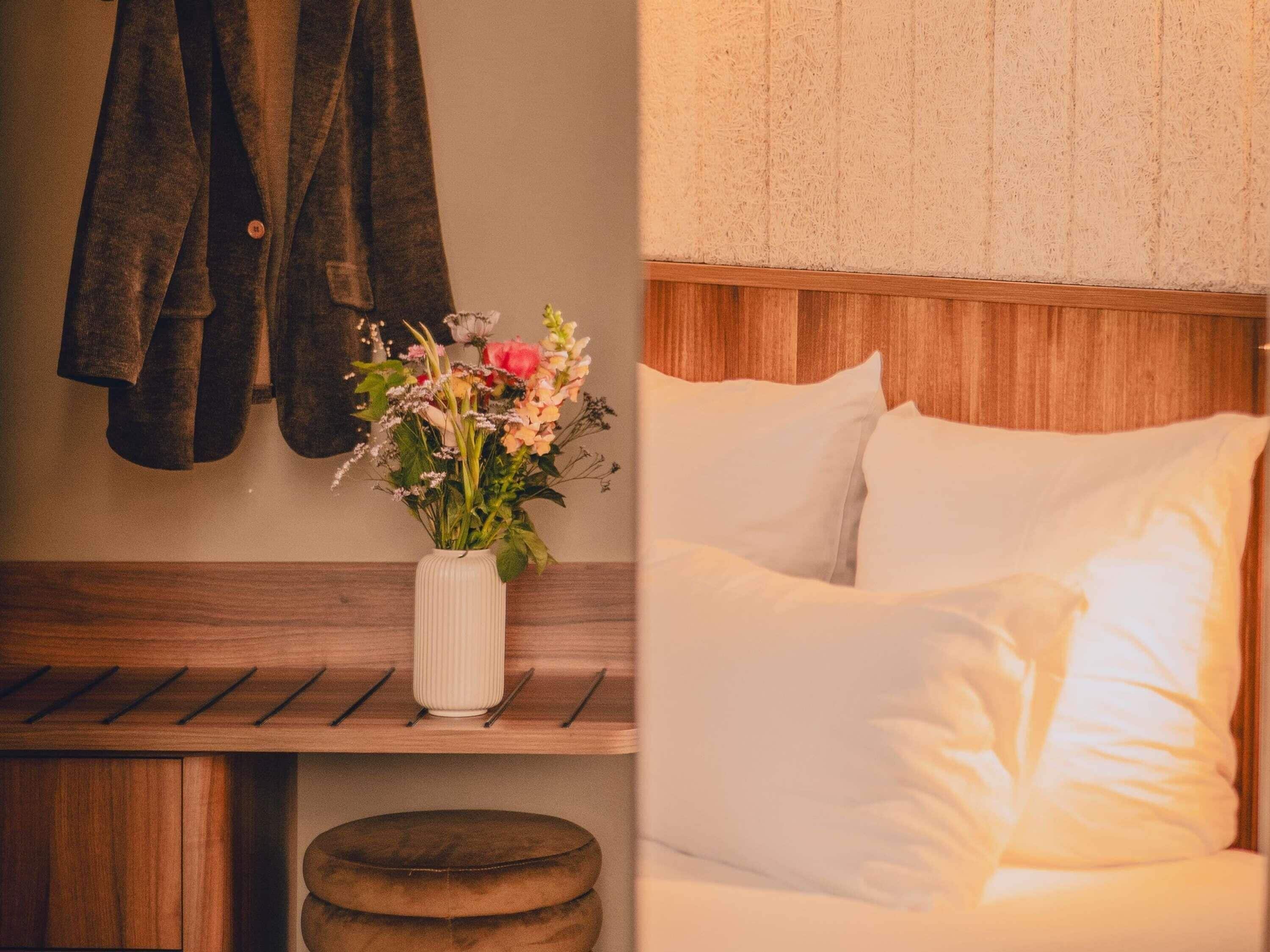 Une chambre avec une tête de lit en bois, un lit blanc avec des oreillers, un banc en bois, un vase blanc avec des fleurs et une veste suspendue.