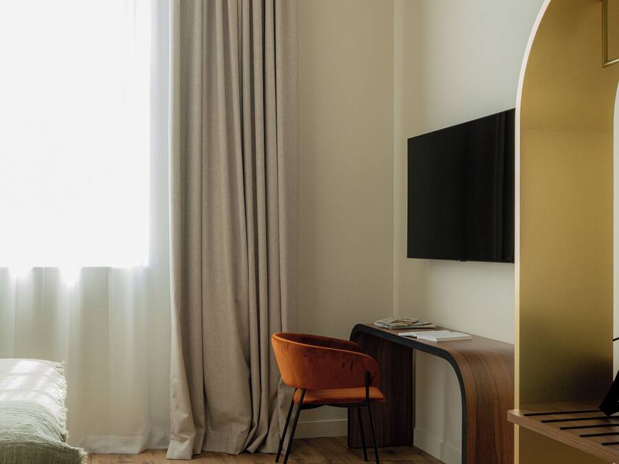 Une chambre moderne avec un bureau en bois, un téléviseur fixé au mur et une grande fenêtre avec des rideaux.