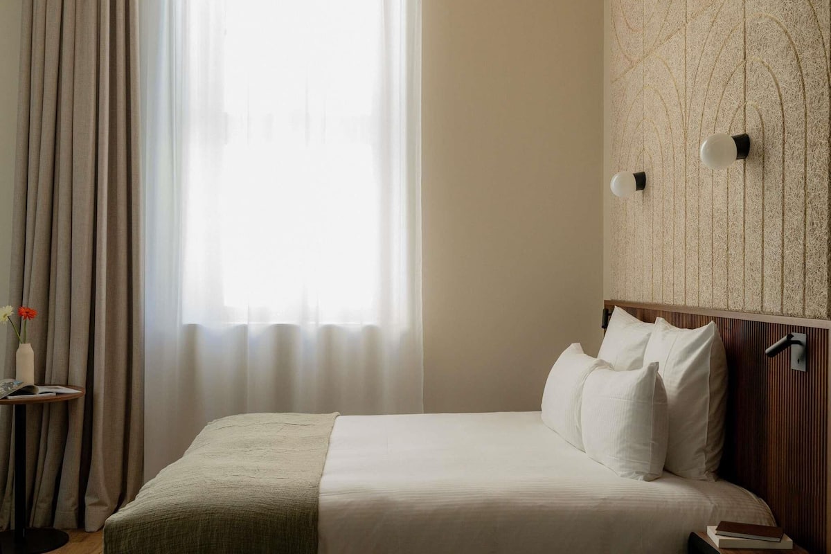 A neatly made bed with white linens, a bedside table with a book, and a wall-mounted light fixture.