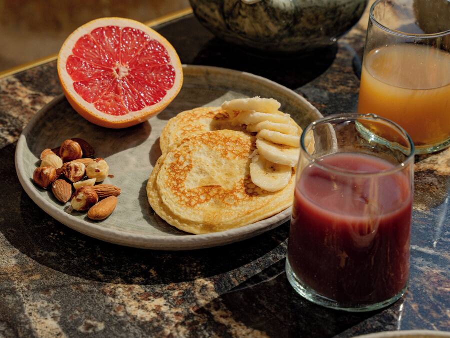A breakfast table with a plate of pancakes, sliced bananas, and nuts, a glass of orange juice, and a teapot.