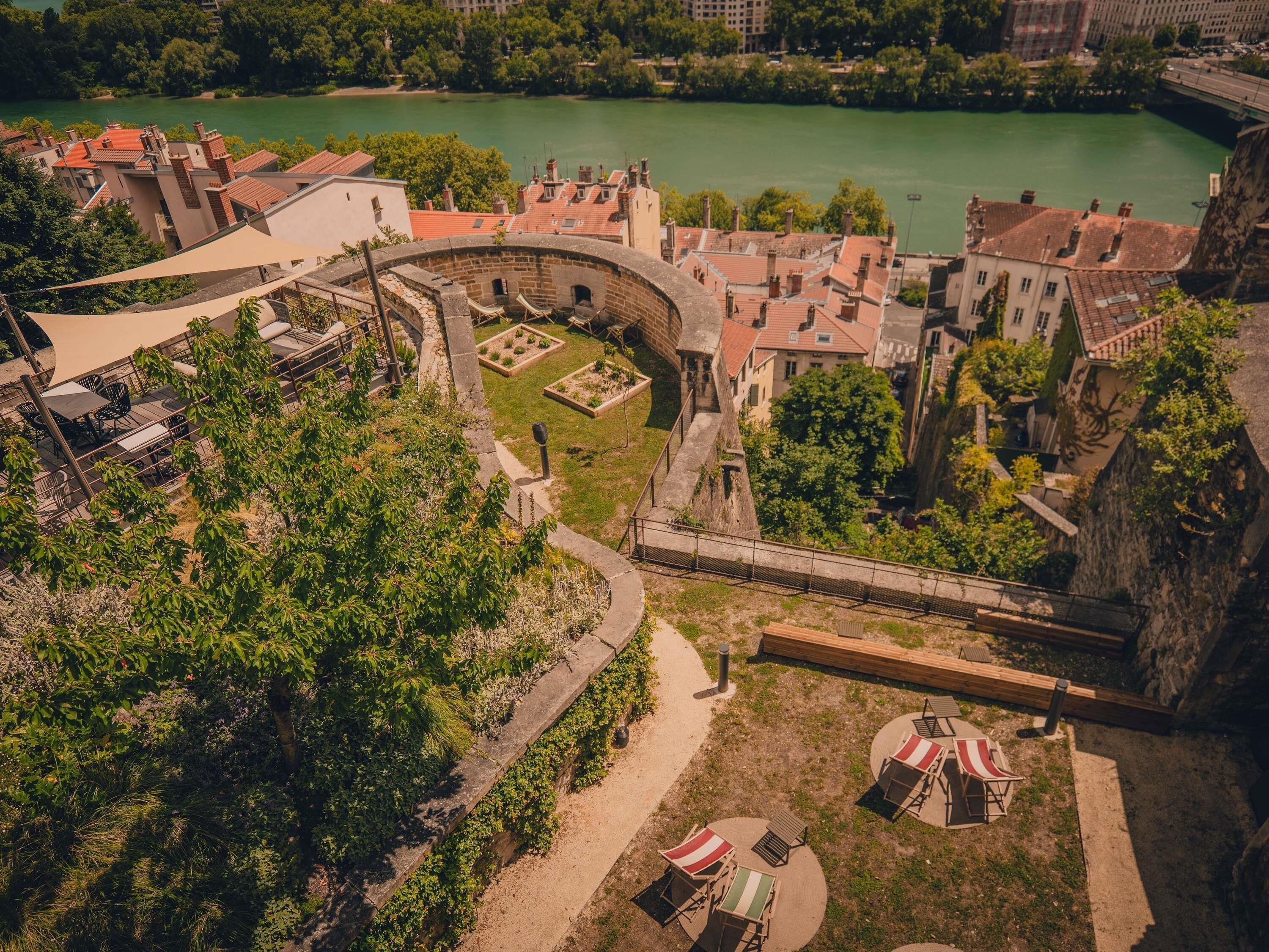Vista aèria d'un jaciment històric amb una estructura de pedra circular, un riu al fons i un passeig pavimentat amb zones de seure.