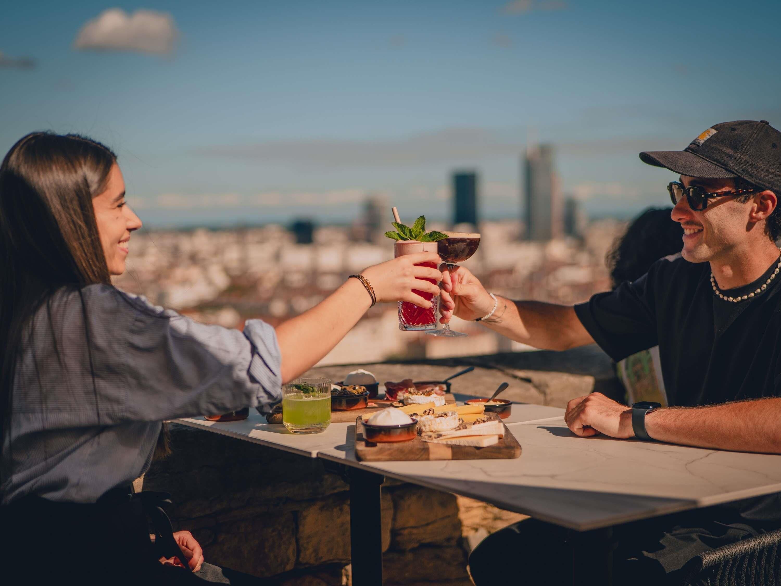 Dos personas brindando con bebidas en una mesa al aire libre con vistas a la ciudad.