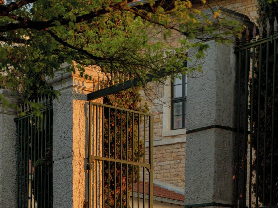 A gated entrance with a metal gate, a stone wall, and a building with a tiled roof.