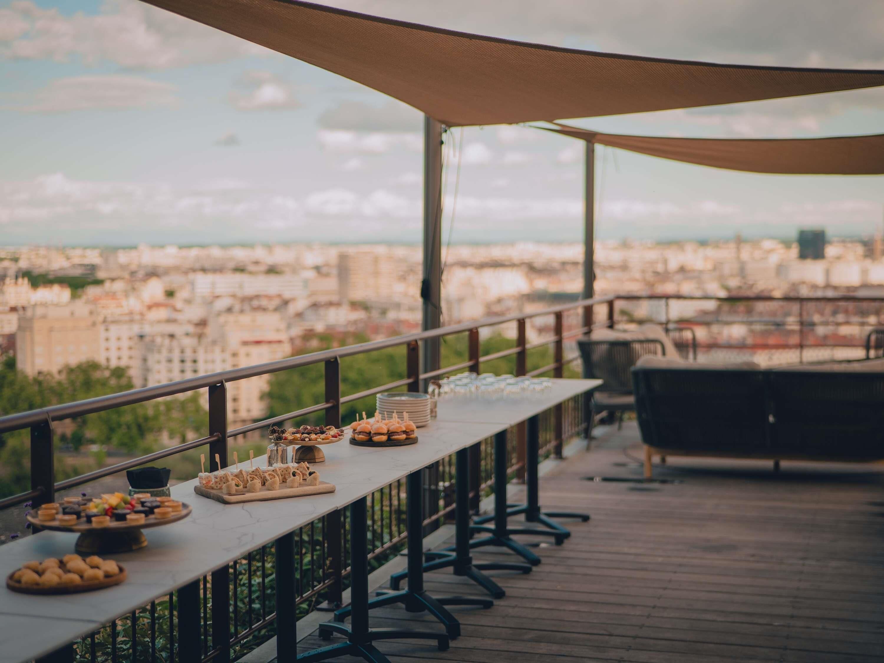 An outdoor terrace with a city view, a long table with food and drinks, and a grill area.