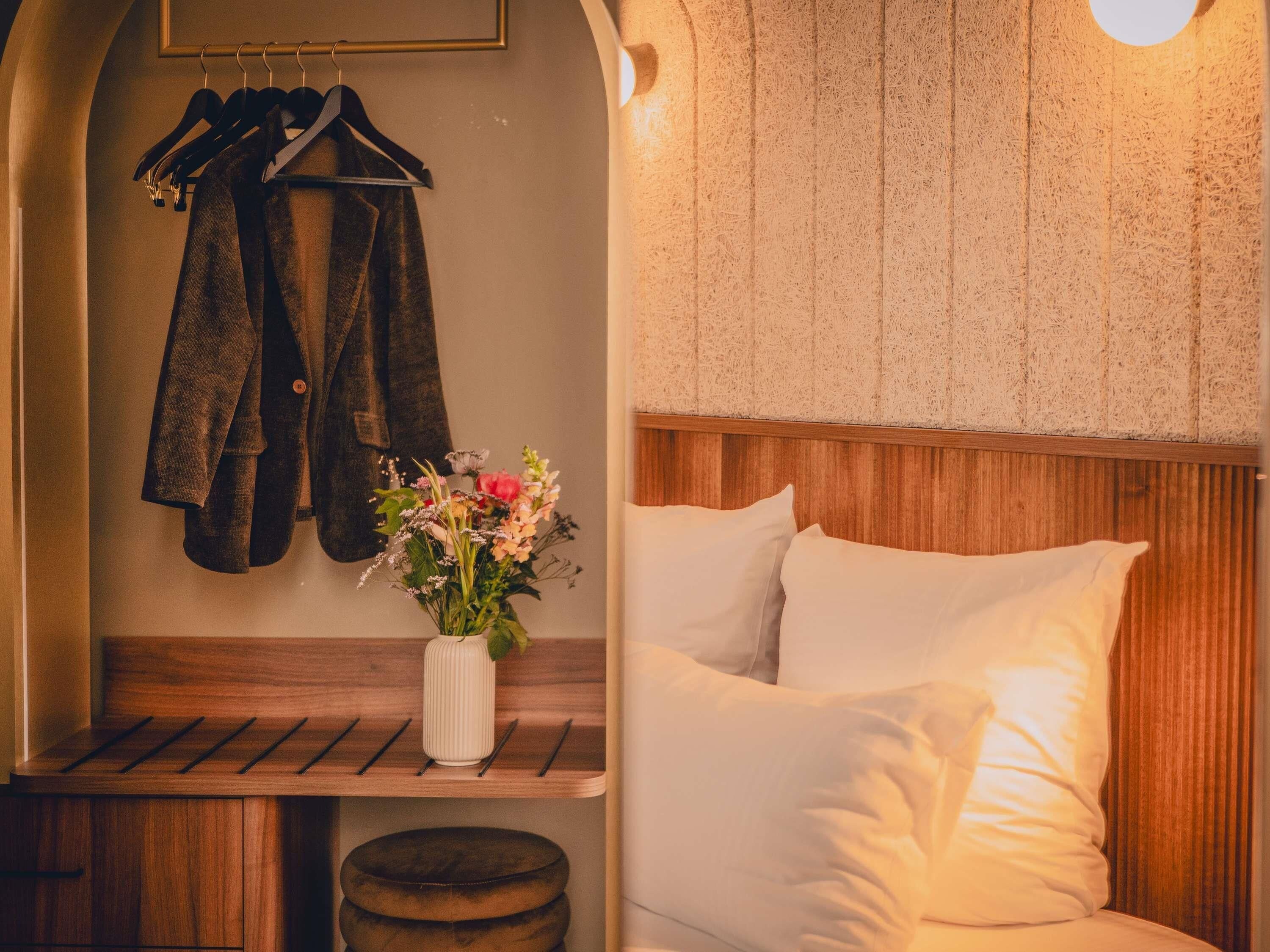 Un dormitorio con cabecera de madera, cama con ropa de cama blanca, un banco con un florero y un perchero con ropa colgada.