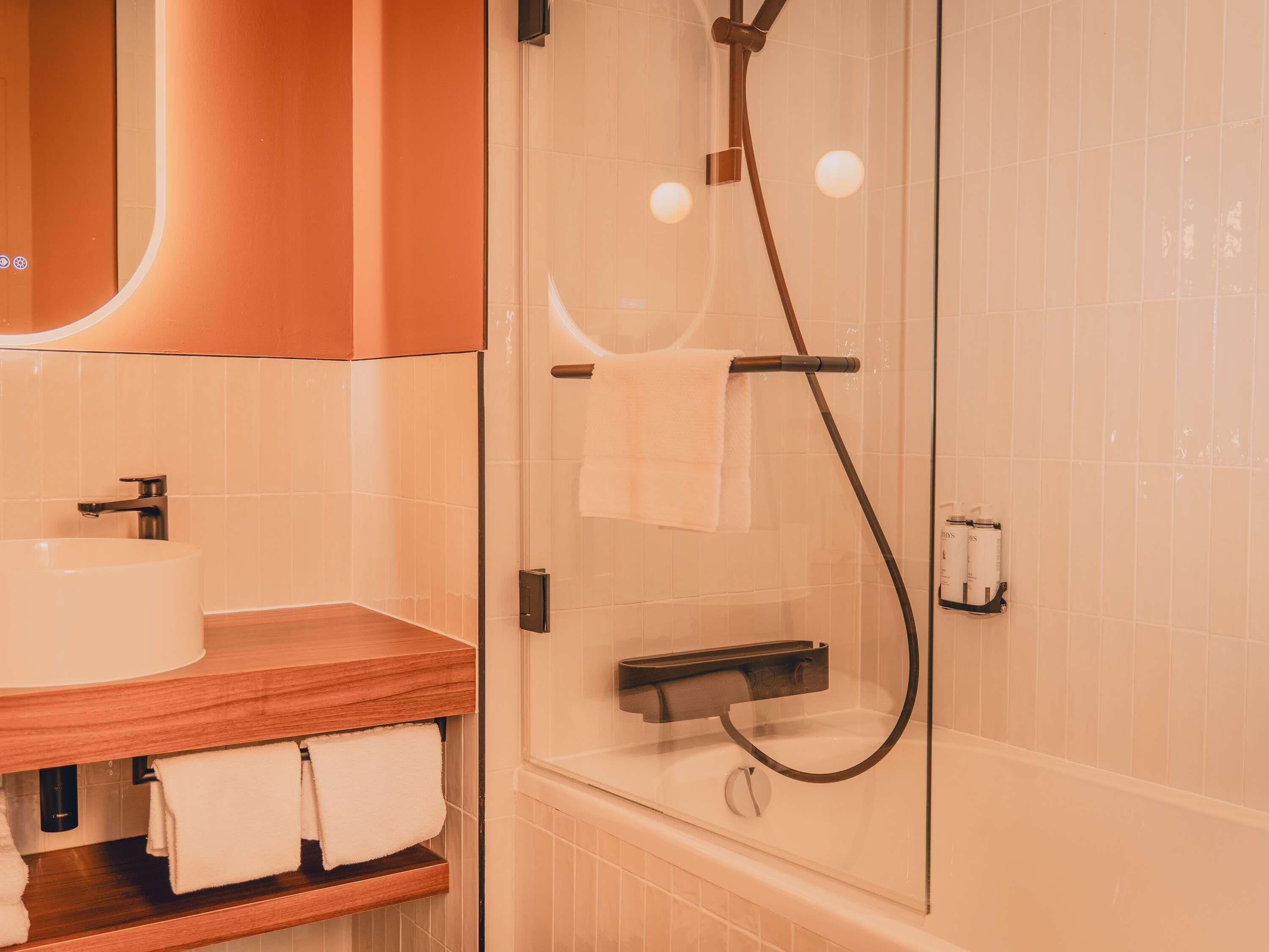 Un baño con ducha envuelta en vidrio, un tocador de madera con lavamanos y un espejo sobre el tocador.