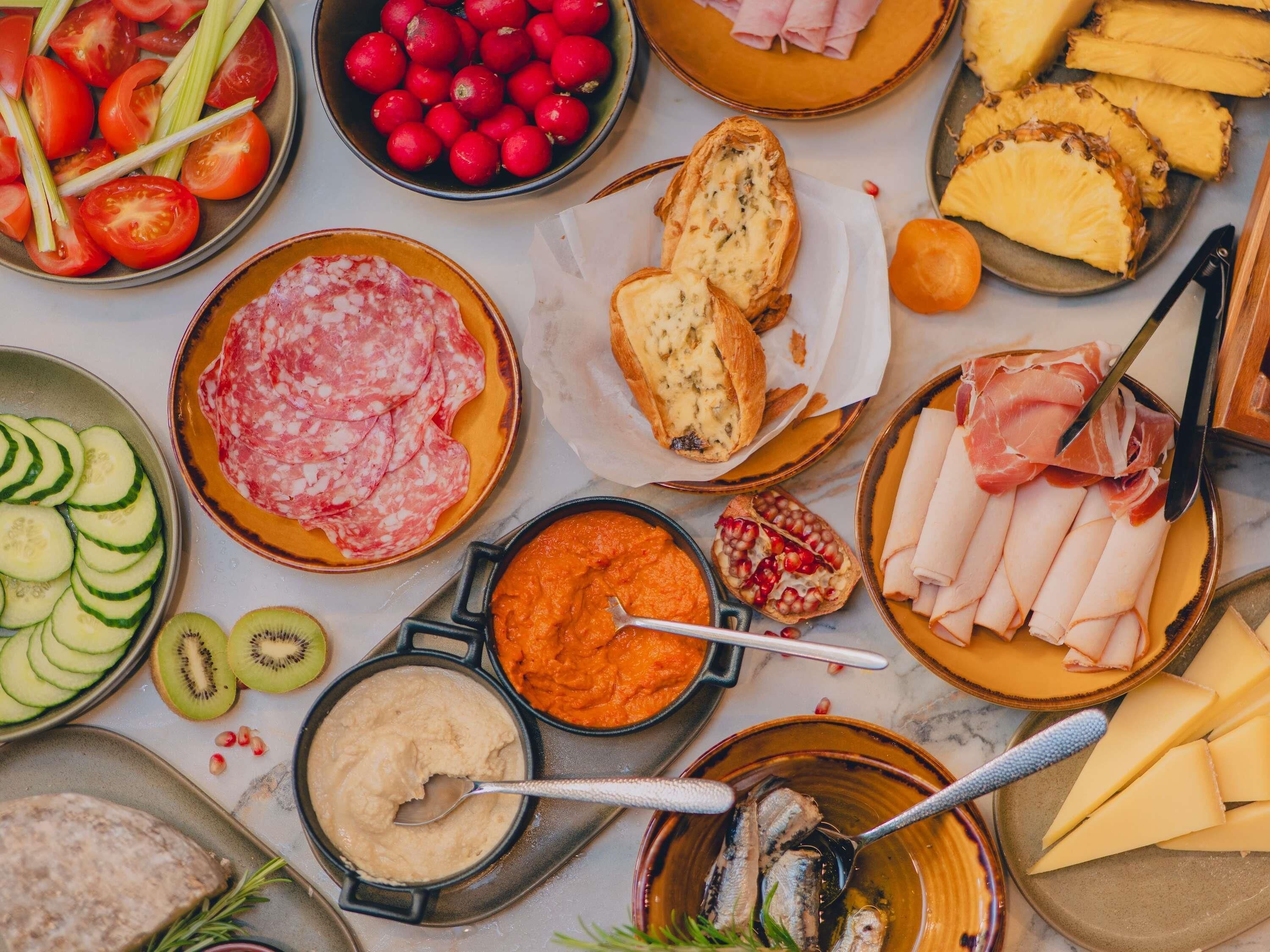 A variety of food items including sliced tomatoes, cucumbers, kiwi, pomegranate seeds, and different types of cheese and cured meats.