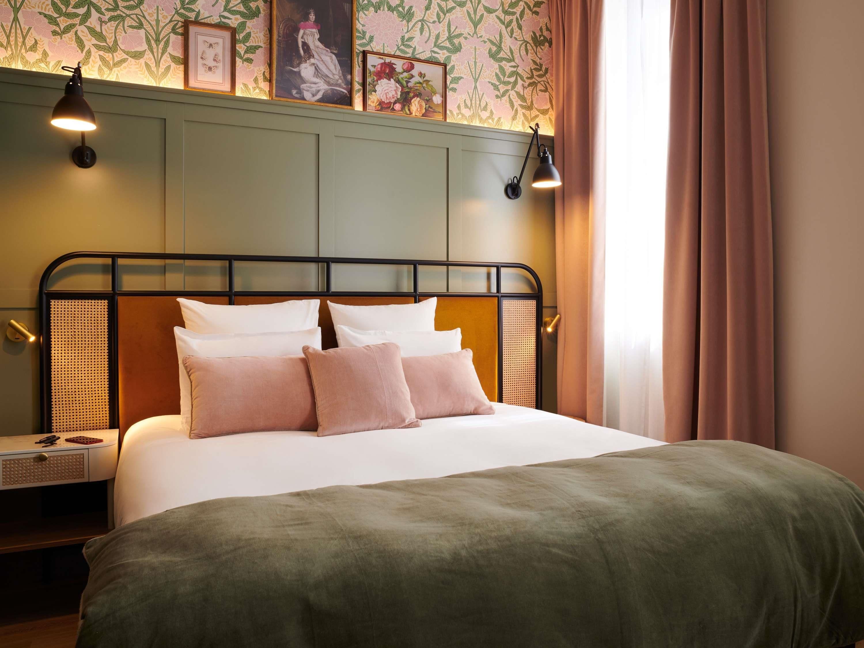 Una cama bien hecha con una colcha verde y varias almohadas, una lámpara de pared y un cuadro enmarcado sobre la cama.