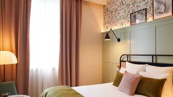 Ein Hotelzimmer mit Bett, Tisch mit Getränk und Speisekarte, Vorhängen und Wand mit floralem Wandpapier.