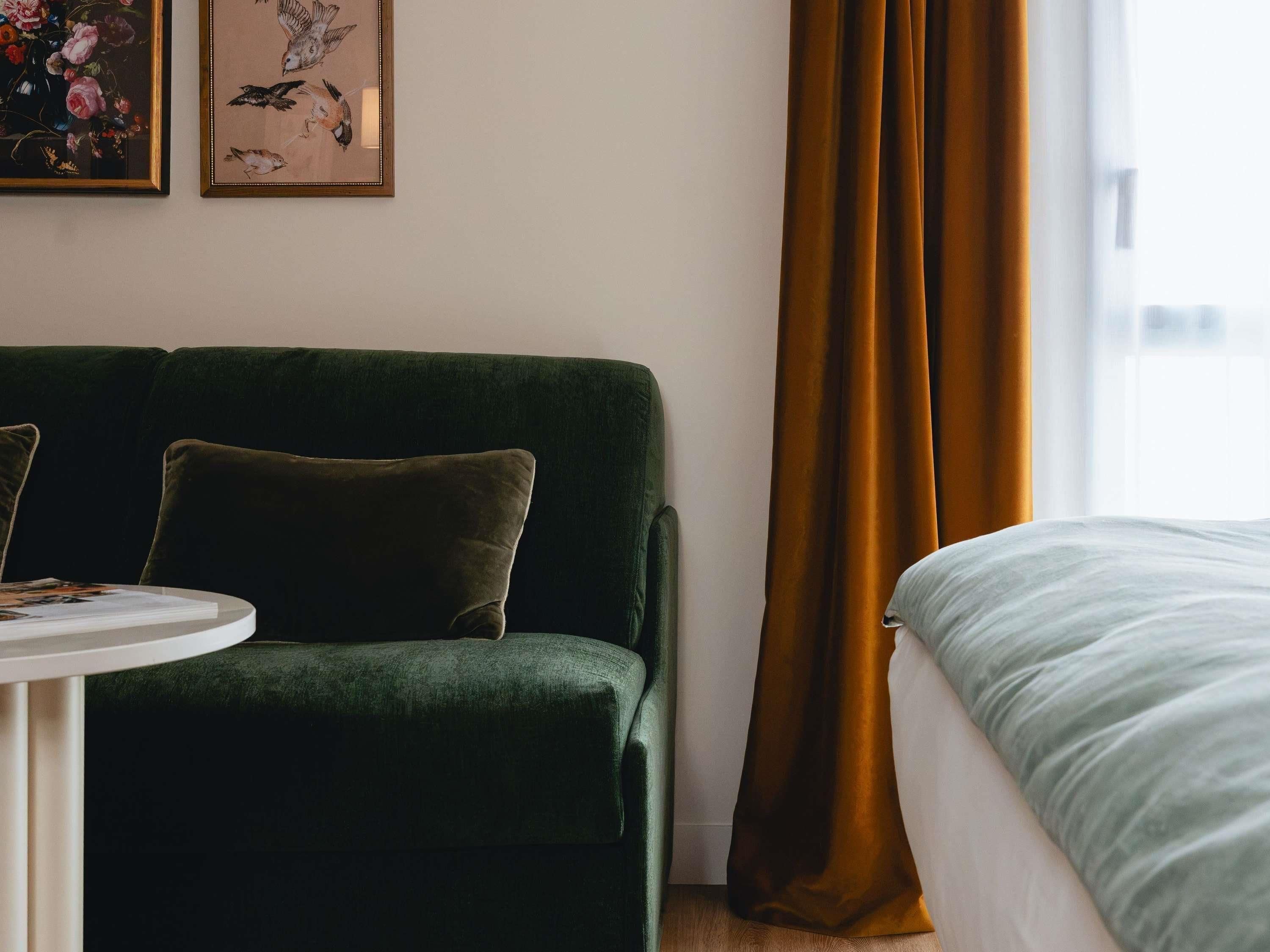 A room with a green sofa, a small table with a magazine, and a bed with a white comforter.