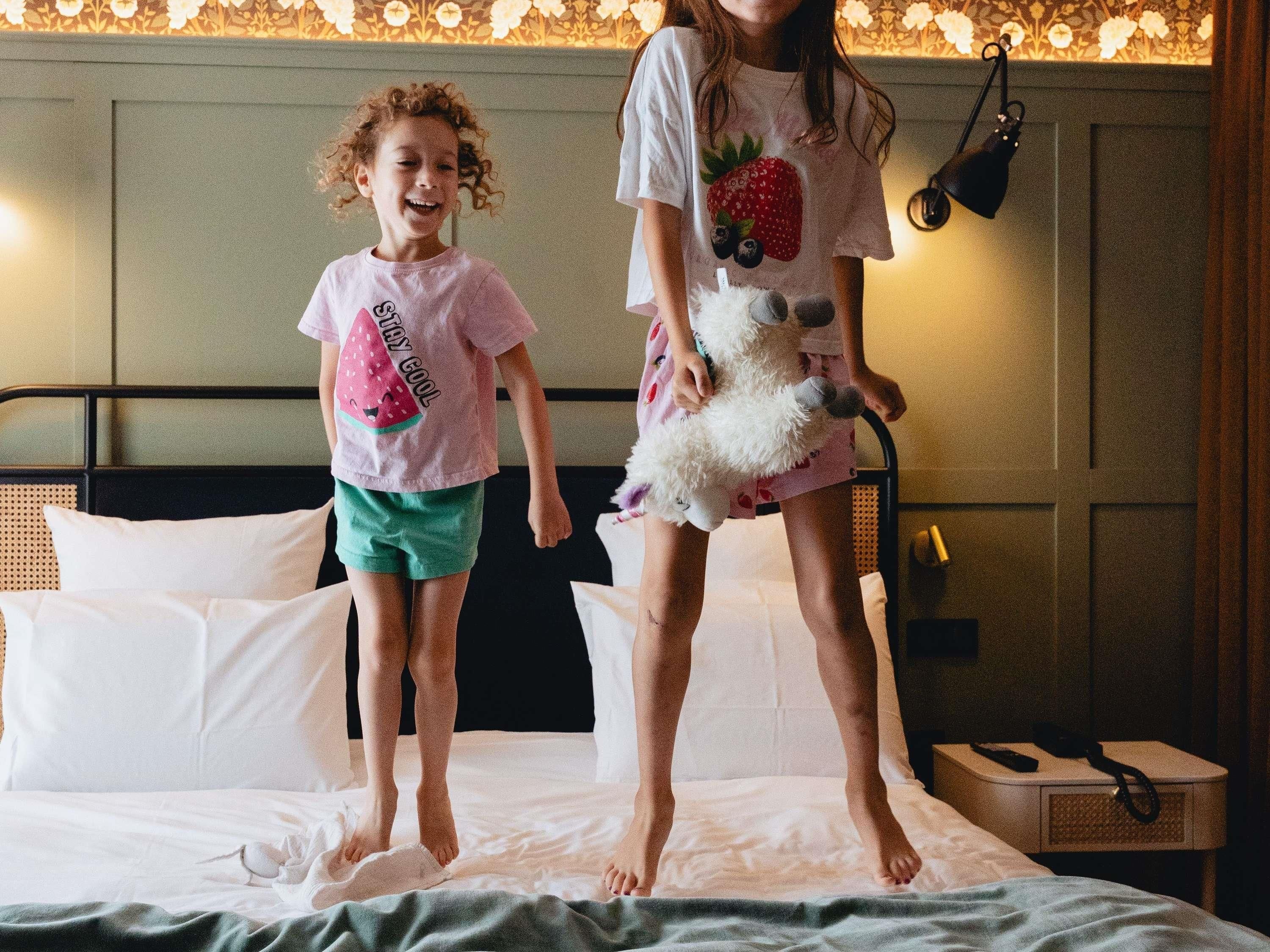 Deux enfants sautent sur un lit dans une pièce dont le papier peint est à motifs, où se trouvent une lampe de chevet et une table de nuit avec un téléphone.