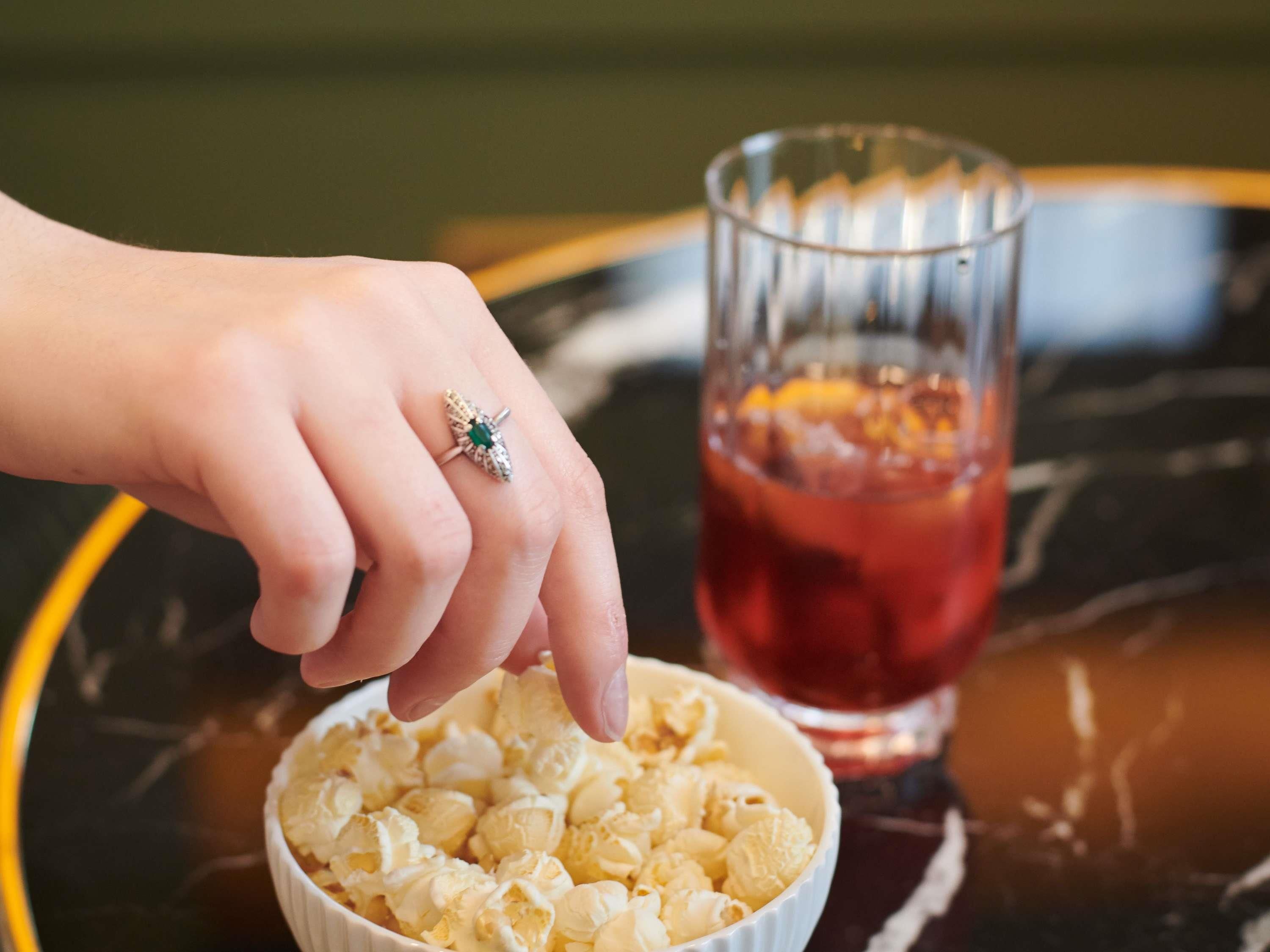 Una mano alcanzando palomitas de maíz de un bol, junto a un vaso de bebida roja y un anillo sobre la mesa.