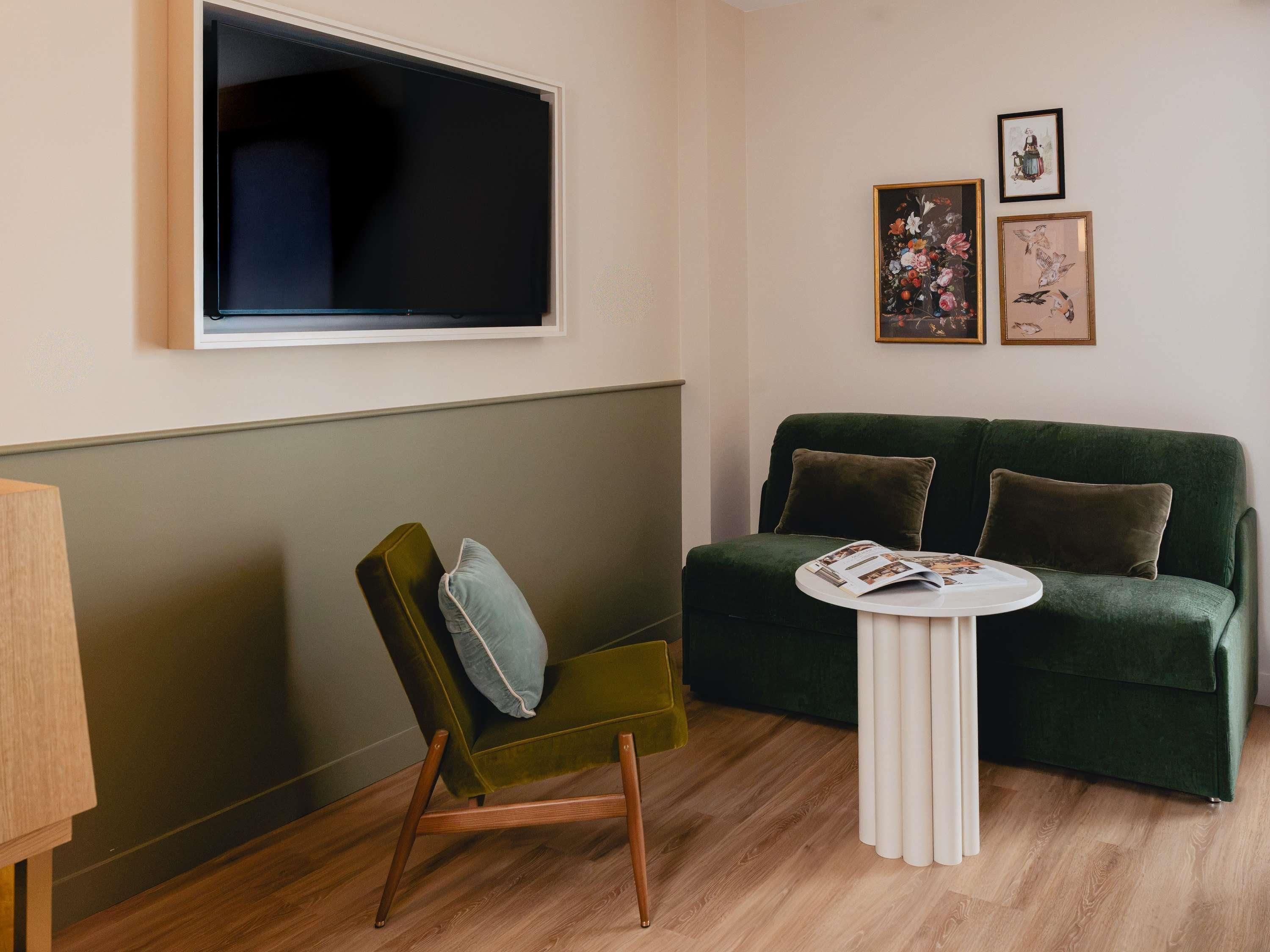 A room with a green sofa, a green armchair, a white side table, and a flat-screen TV mounted on the wall.