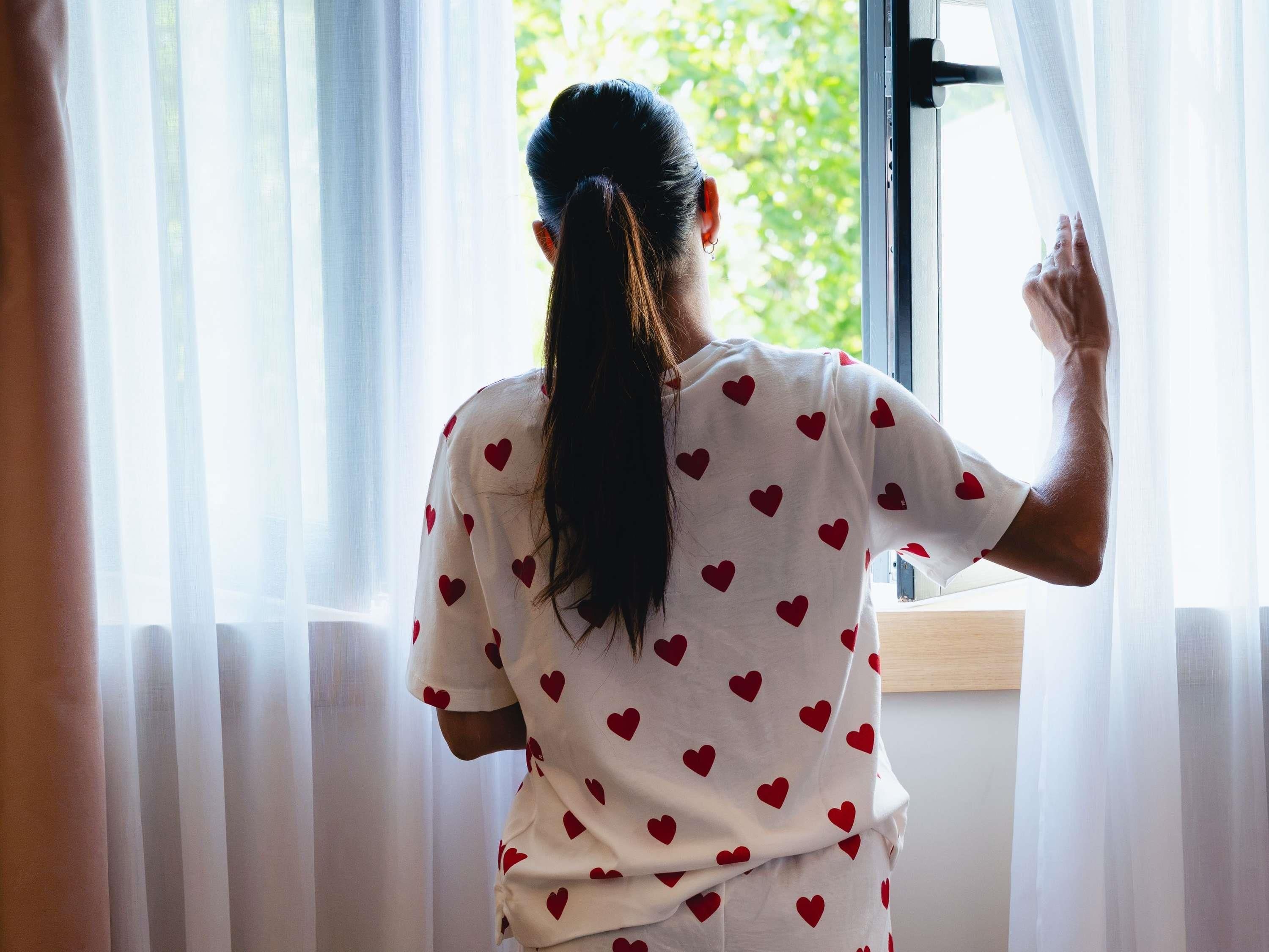 Una persona de pie junto a una ventana con cortinas translúcidas, mirando hacia el exterior.