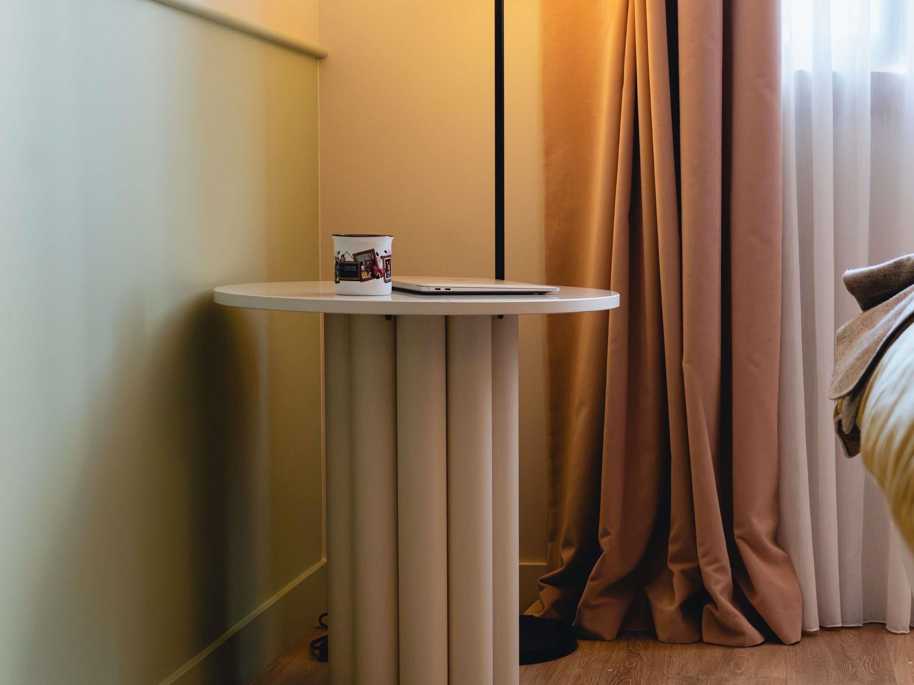 A small round table with a mug and a smartphone, a bed with a blanket, and a window with curtains.