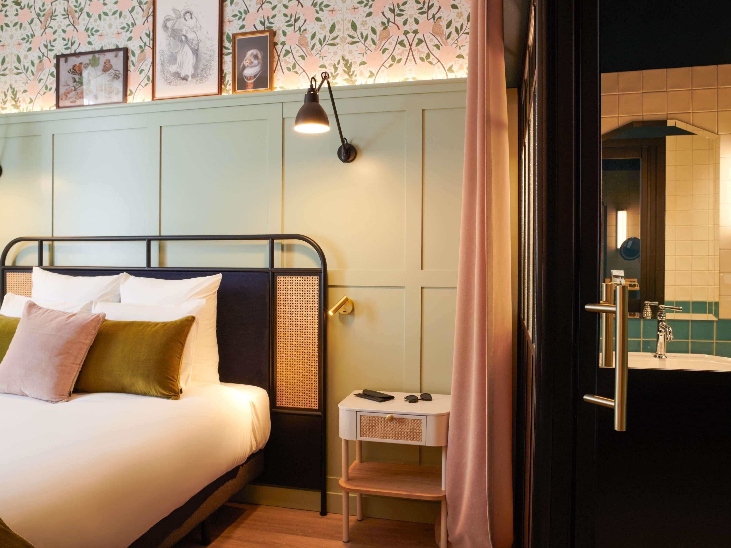 A neatly made bed with pillows, a bedside table with a lamp, a small stool, and a bathroom visible through an open door.