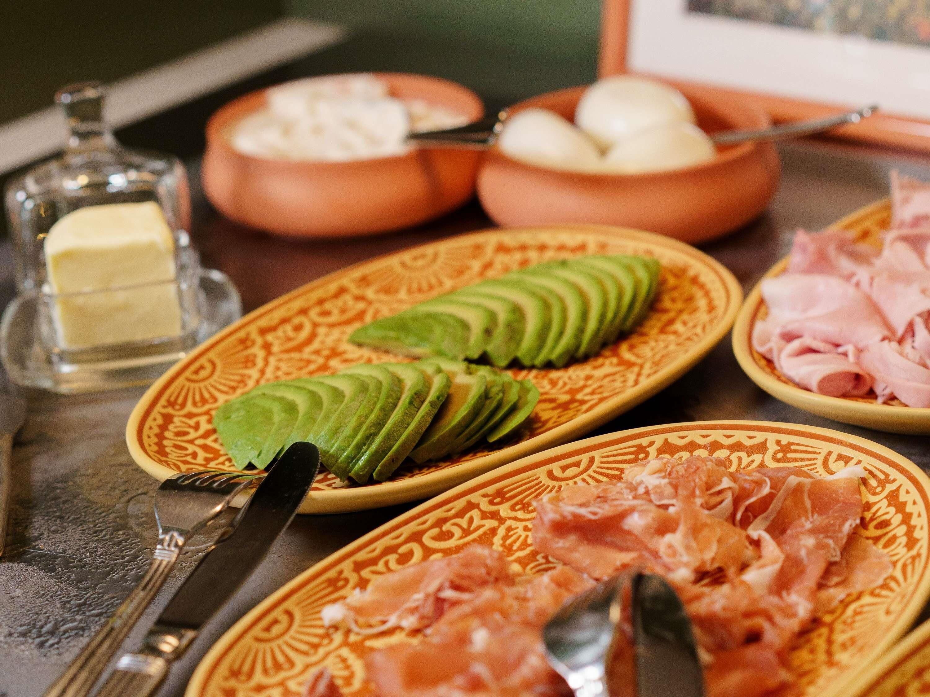 Una mesa con diferentes alimentos, incluyendo aguacate en rodajas, jamón y un tazón de crema blanca.