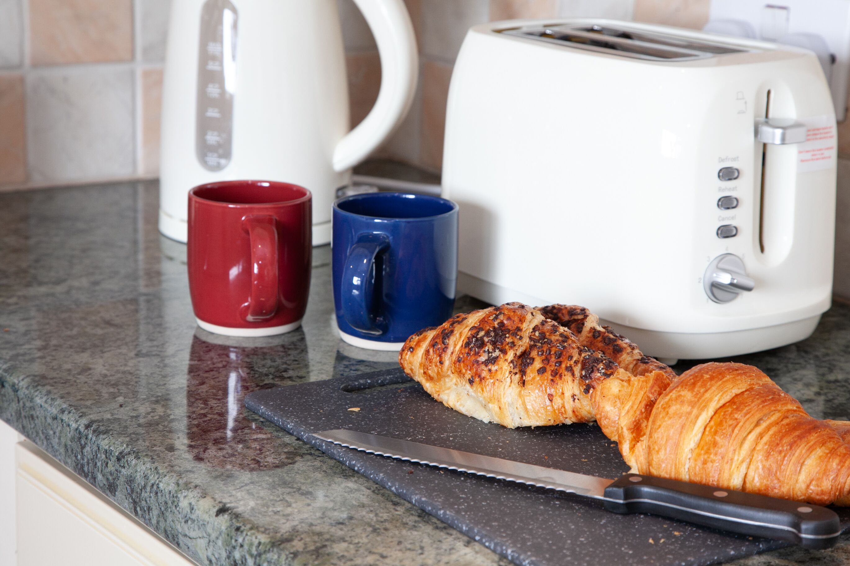Uma bancada de cozinha com uma torradeira, duas canecas, uma faca e um croissant.