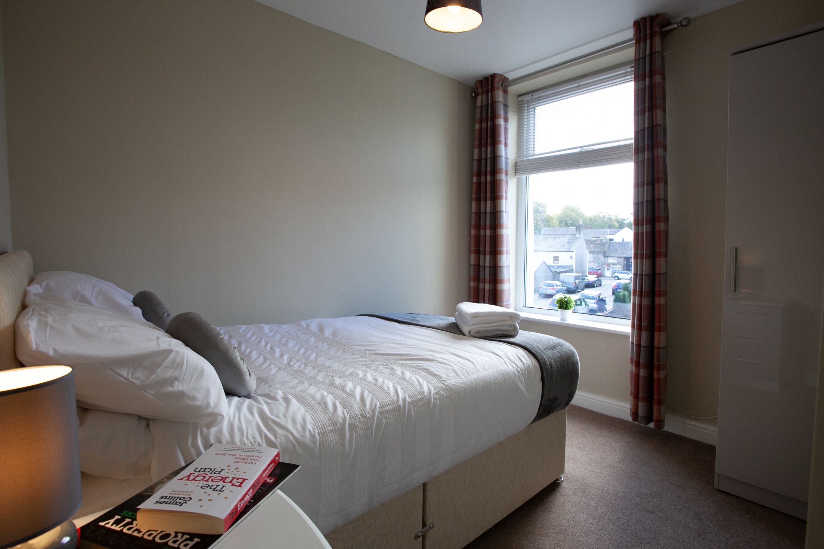 Een slaapkamer met een bed, een nachtkastje met een lamp, een raam met gordijnen en uitzicht op het omliggende landschap.