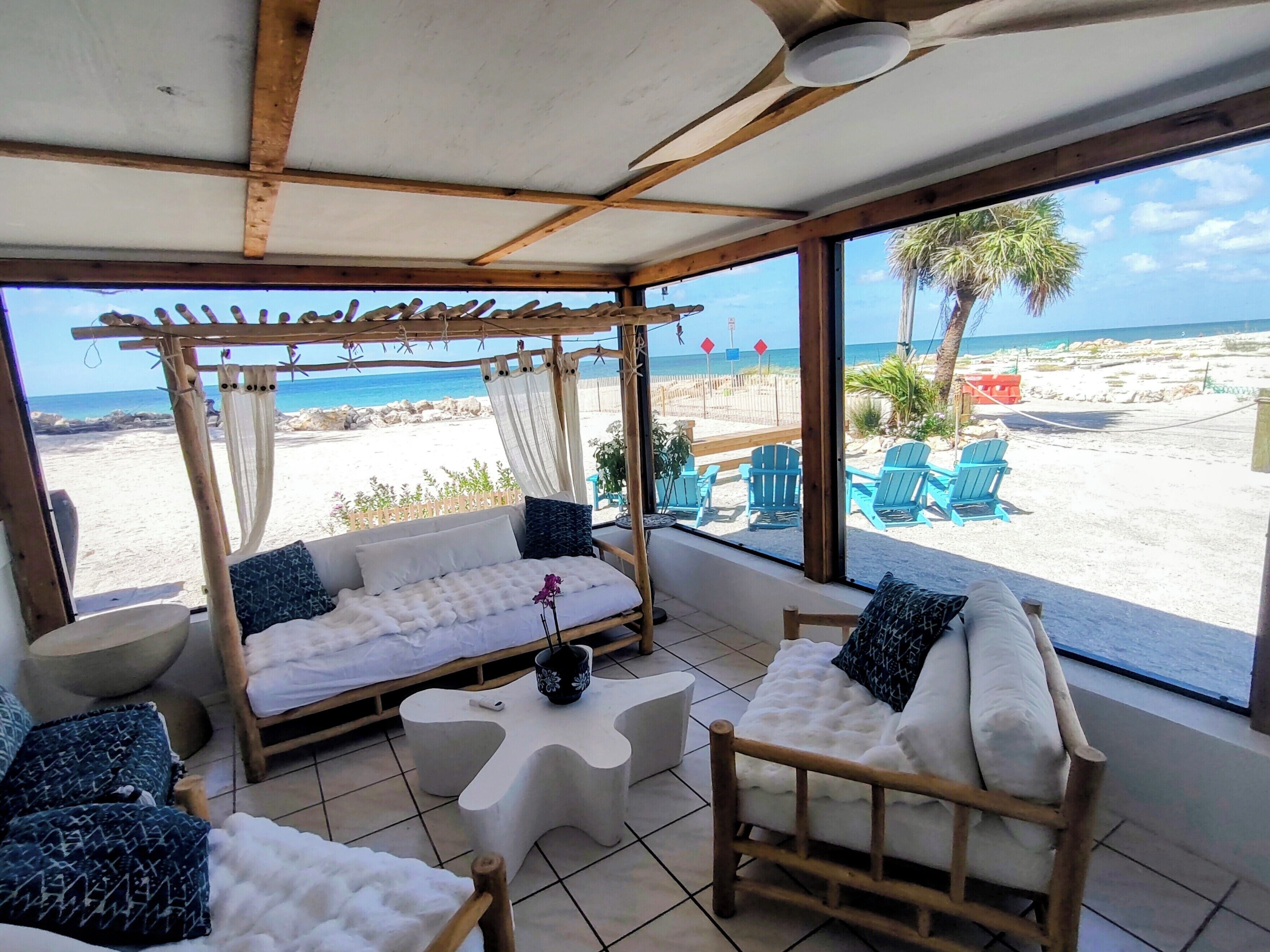A beachside room with a wooden ceiling, a cushioned sofa, a wooden chair, and a view of the beach and ocean.