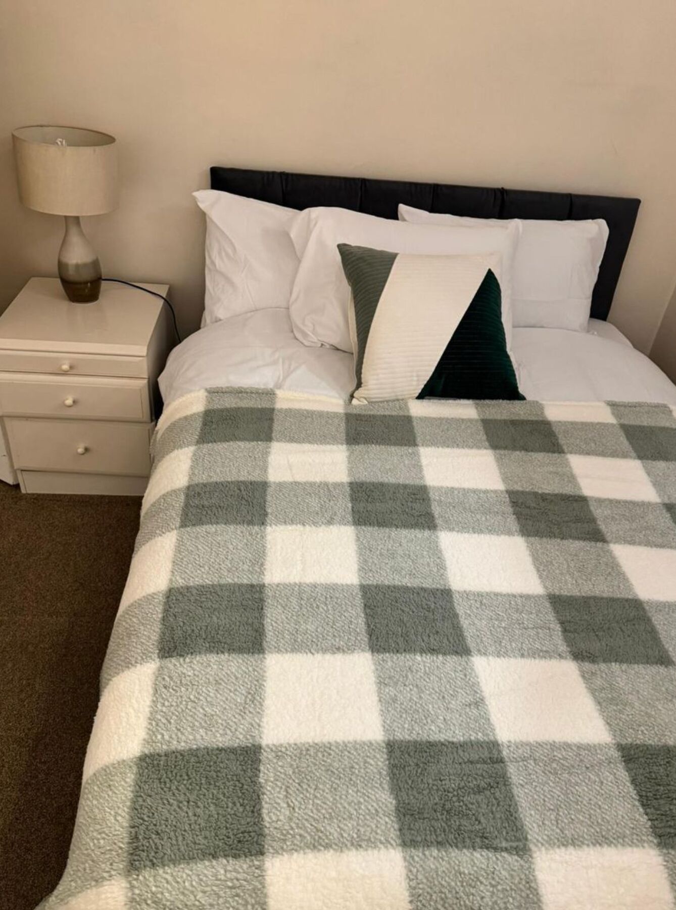 A neatly made bed with a checkered blanket, a bedside lamp, and a nightstand with drawers.