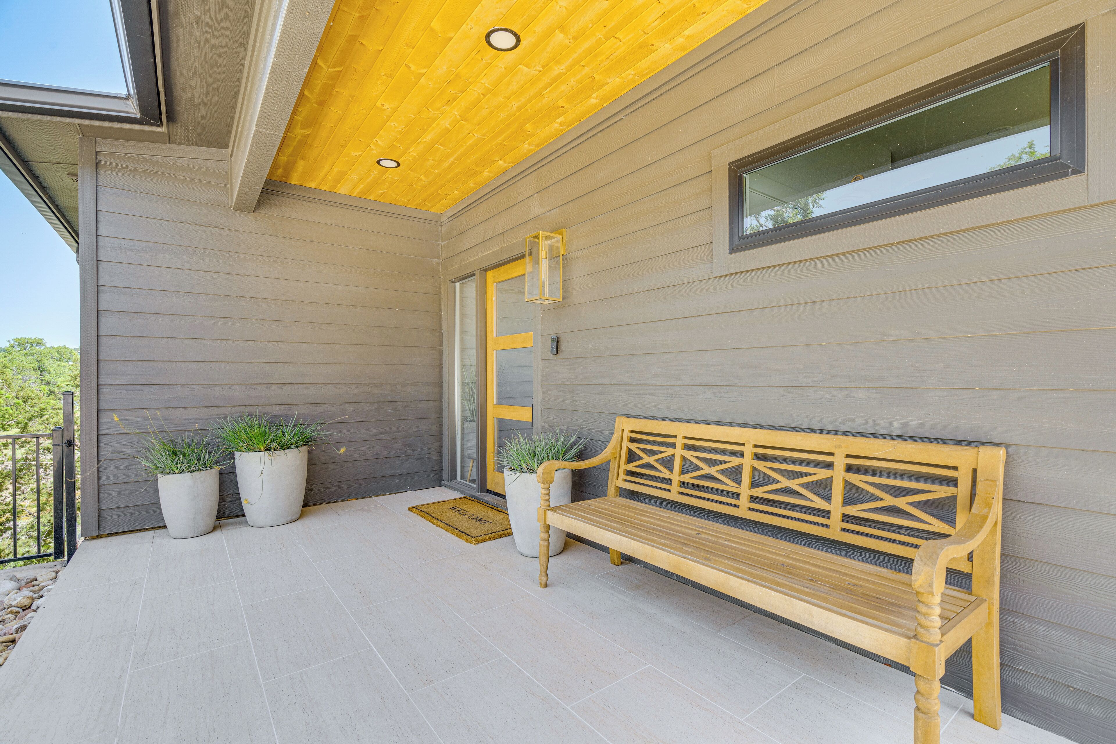 Un banc en bois sur une véranda, avec deux plantes en pot, une porte et une fenêtre.