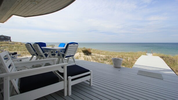 Eine Holzterrasse mit weißen Stühlen und einem Tisch, mit Blick auf einen Strand und einen klaren Meerblick.
