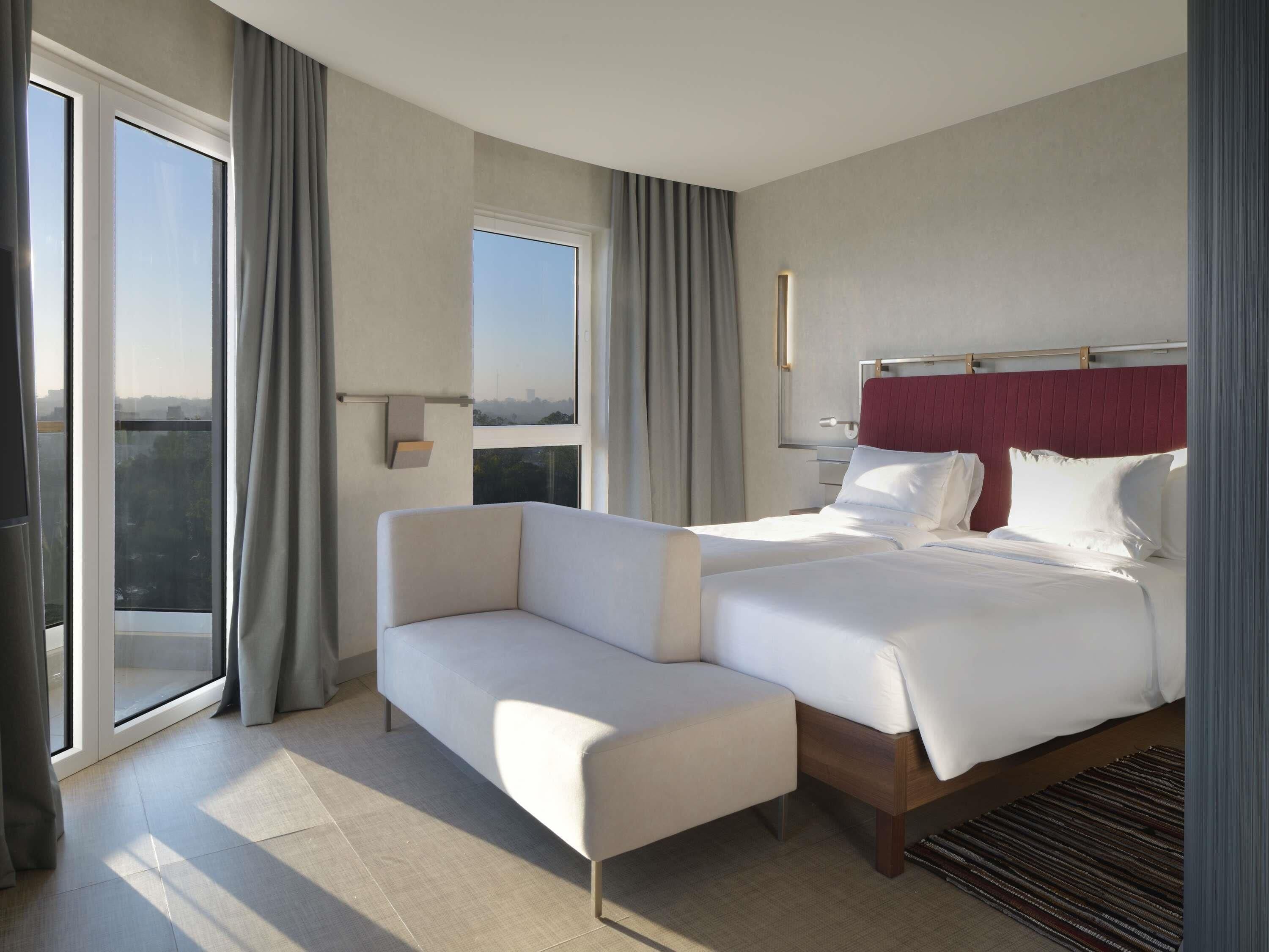Ein Hotelzimmer mit Bett, Sofa, Fenster mit Vorhängen, Nachttisch und Blick auf die Stadt.