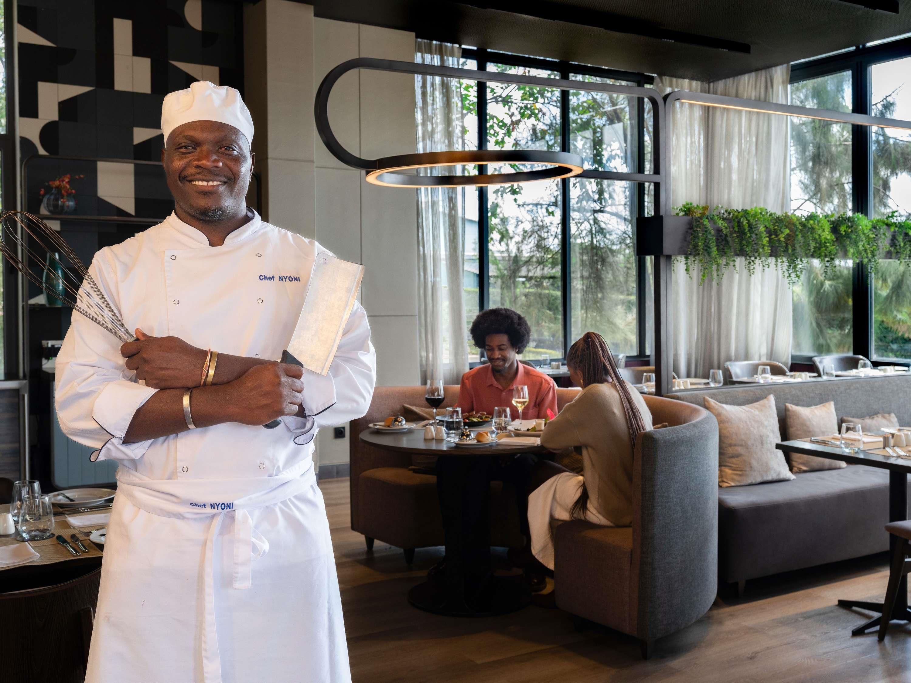 Un chef con uniforme blanco y el texto 'Chef NYONI' en el delantal, de pie en un comedor moderno con un gran ventanal, una mesa redonda con una pareja cenando, y un sofá al fondo.