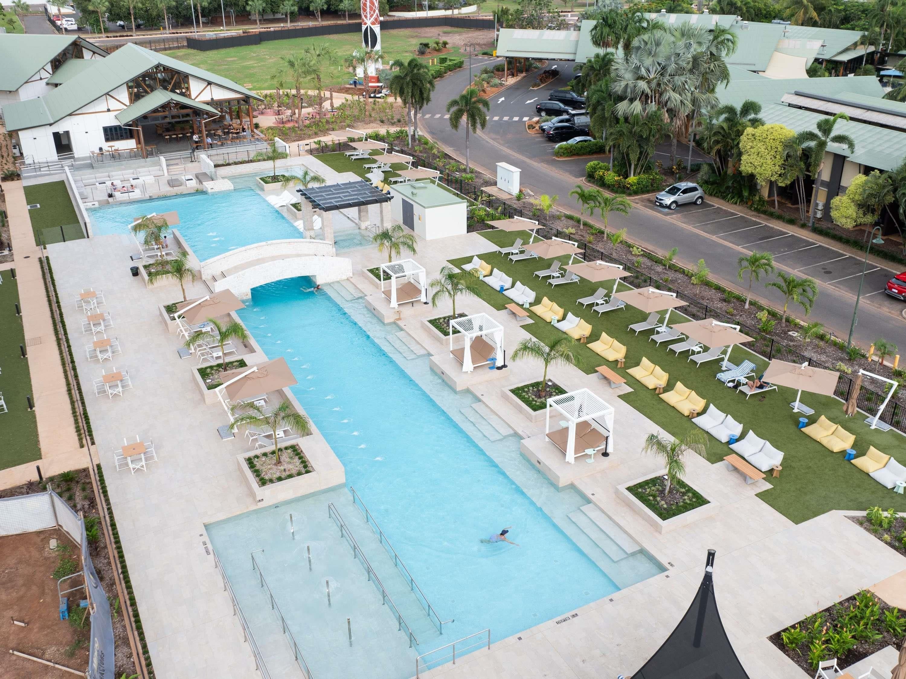 A large swimming pool with multiple lanes, surrounded by lounge areas with chairs and umbrellas, and a parking area with cars.