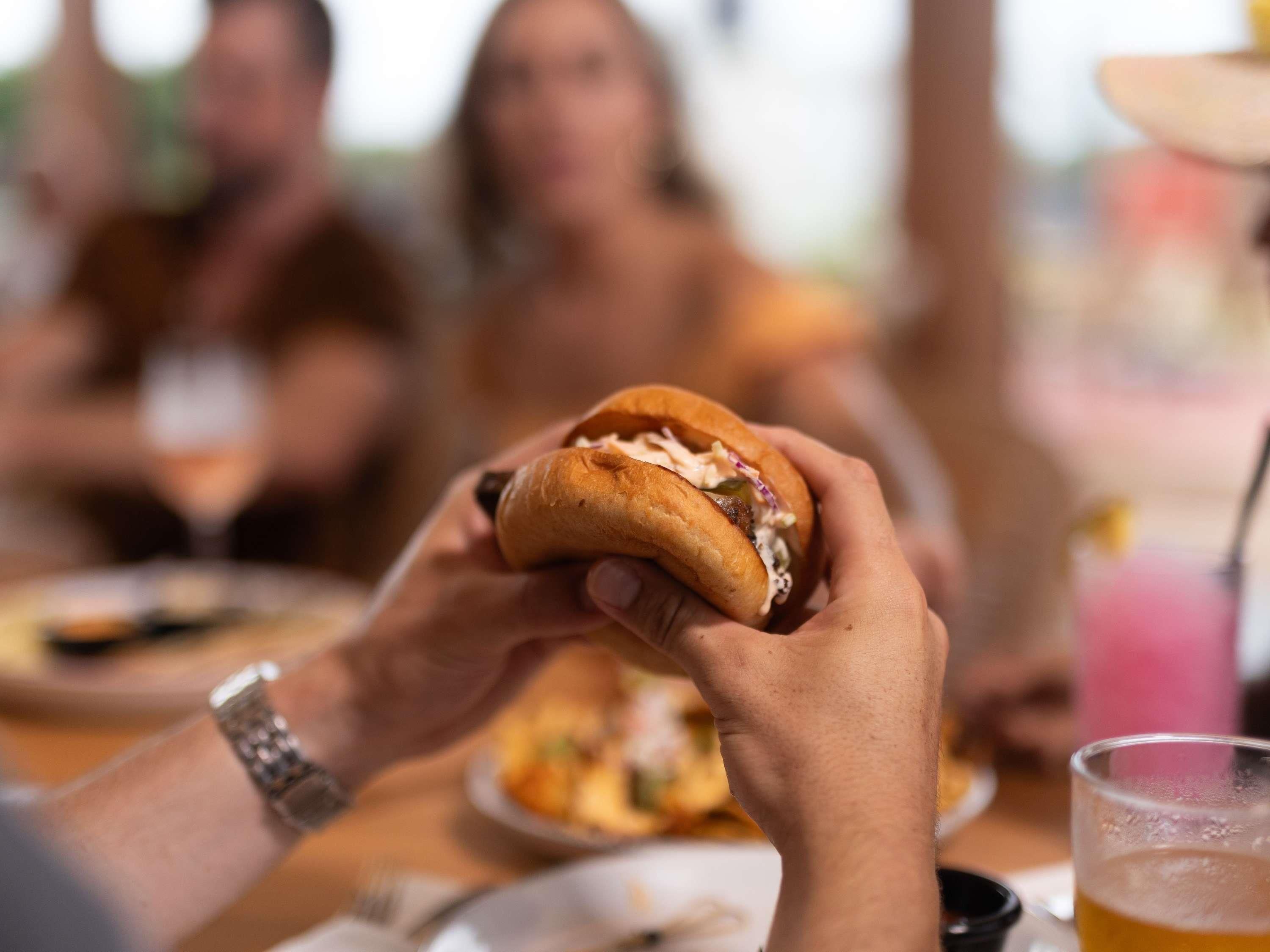 A person holding a sandwich with a bun, with a blurred background of other people.