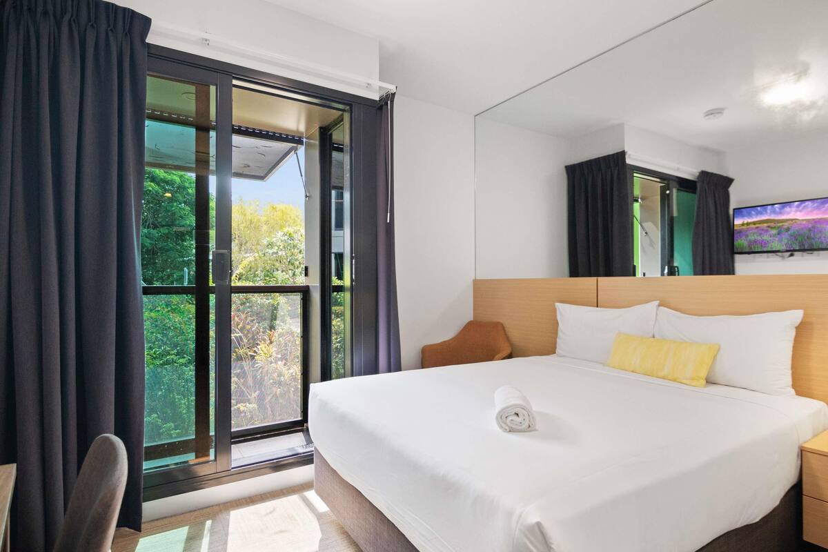A hotel room with a large bed, a flat-screen TV, a window with curtains, and a view of greenery outside.