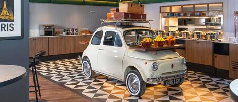 A vintage car parked in a modern kitchen area with geometric patterned flooring, wooden cabinets, and a counter with food items.
