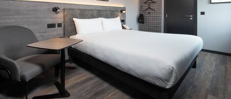 A modern hotel room with a bed, a desk, a chair, and a wardrobe.