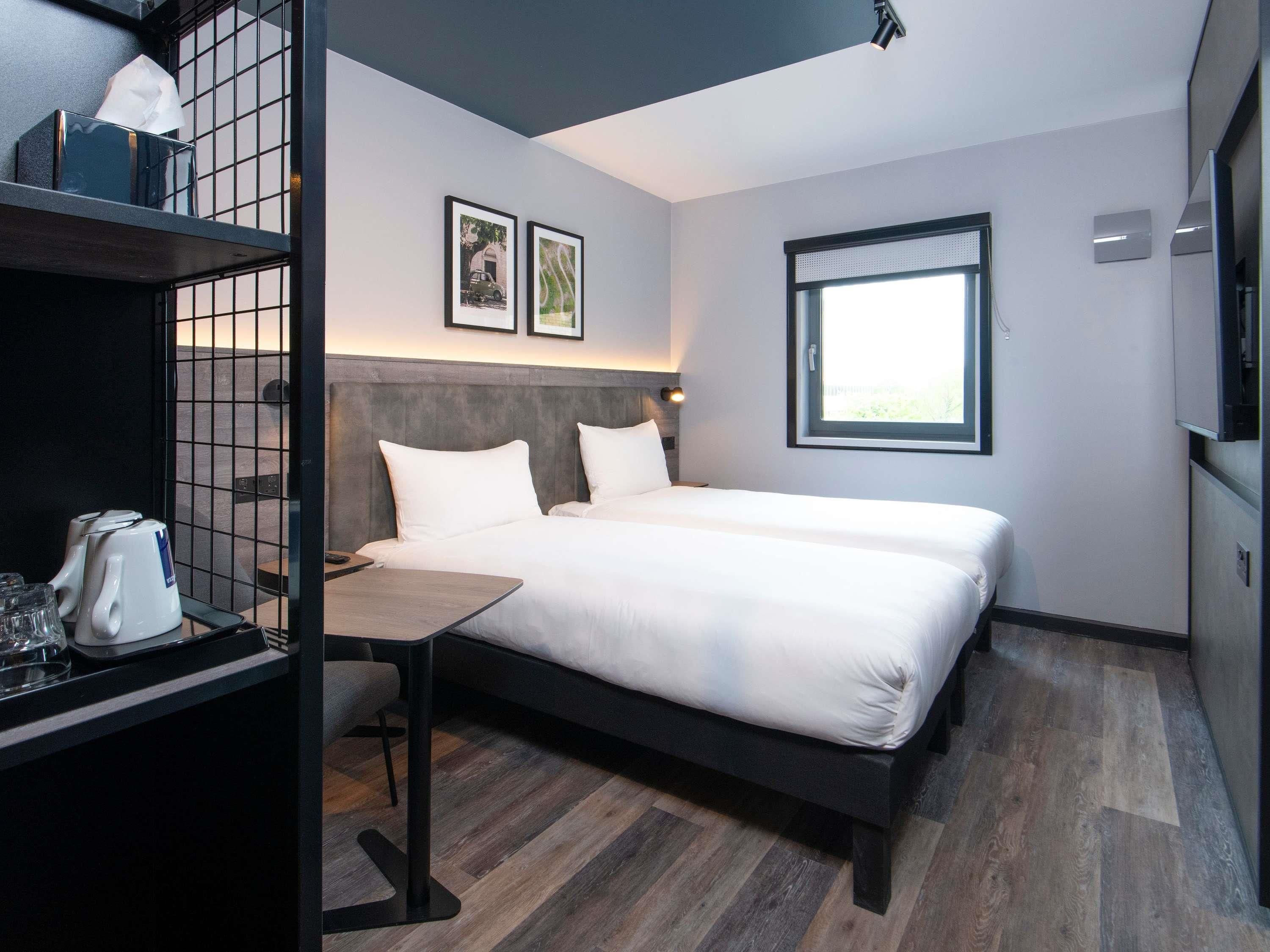 A modern hotel room with a bed, a desk with a kettle, a chair, and a window.