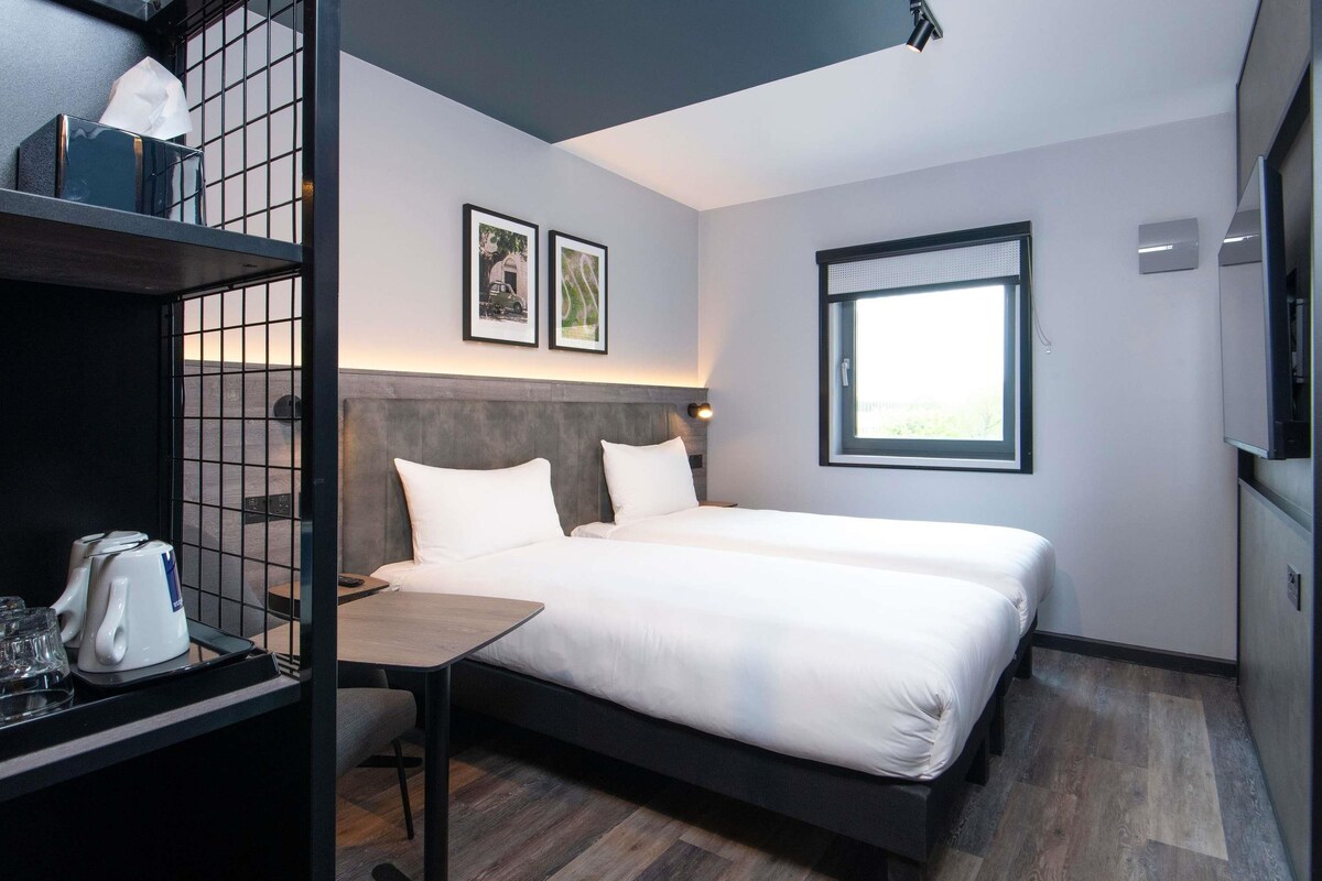 A modern hotel room with a bed, a desk with a kettle, a chair, and a window.