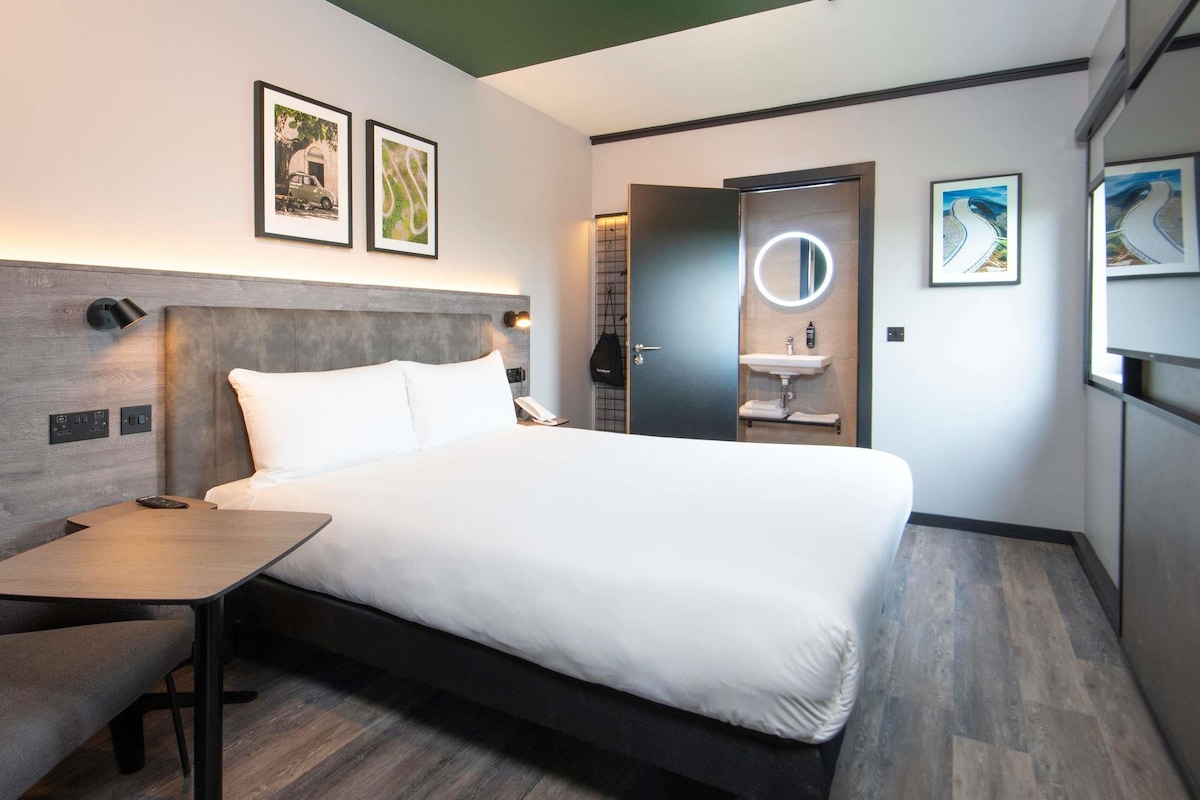 A modern hotel room with a bed, bedside table, and a small desk. There is a bathroom with a mirror and sink.