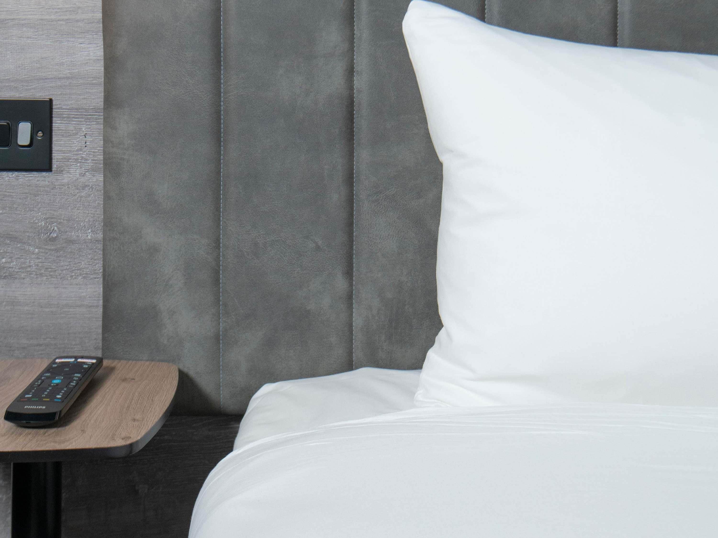 A bed with a white pillow, a remote control on a wooden nightstand, and a grey wall with a switch.
