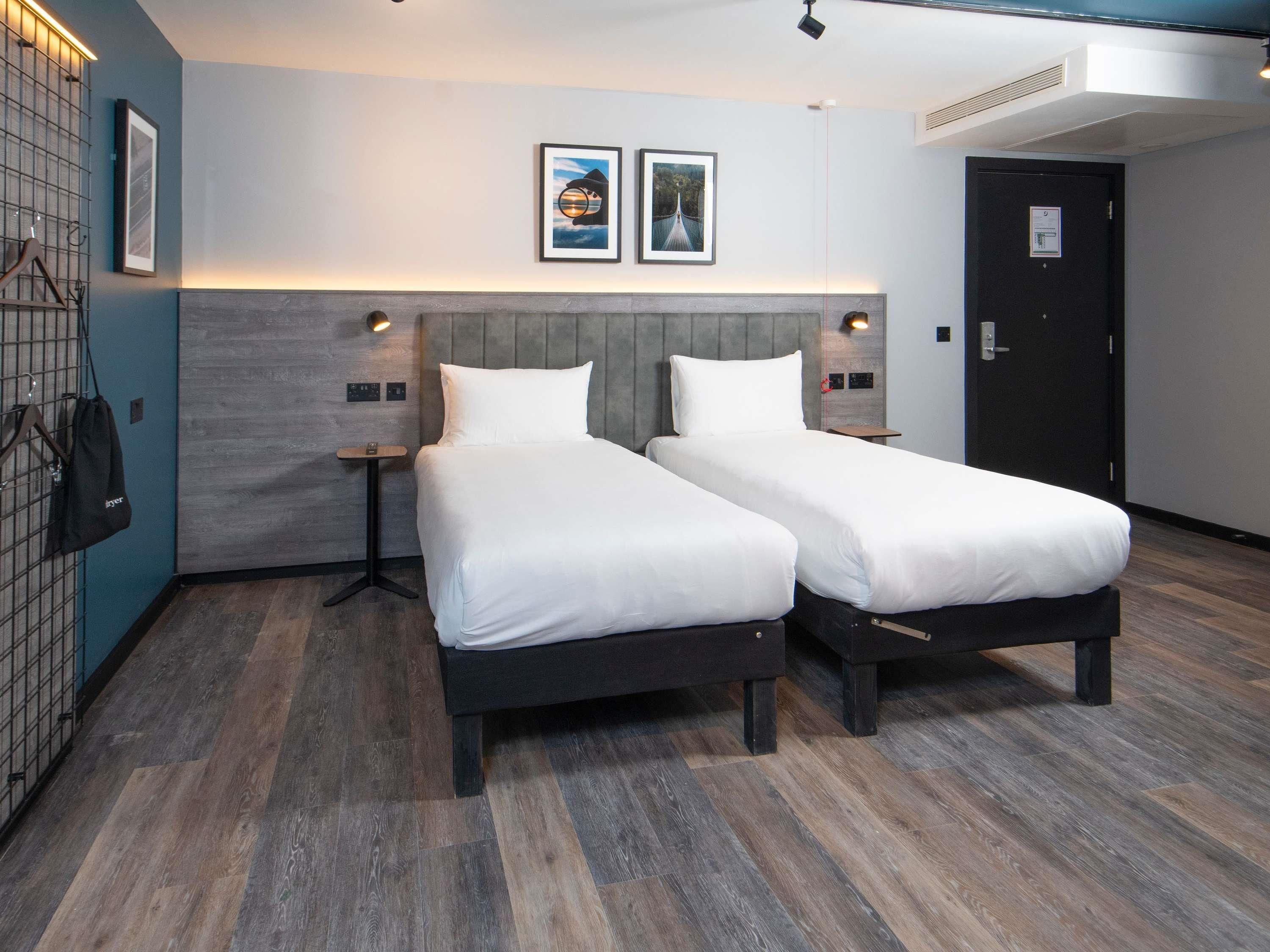 Habitación de hotel con dos camas, una mesita, un estante en la pared y una puerta.