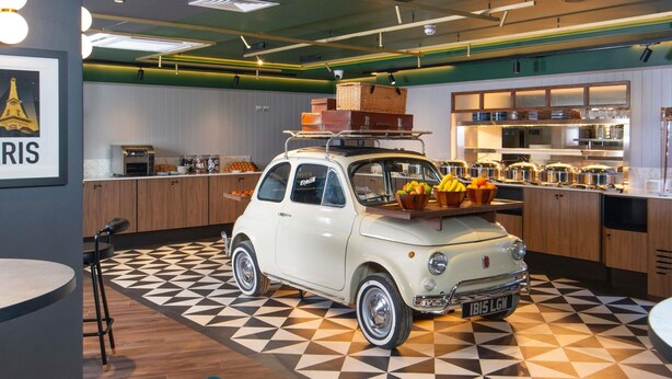 Une voiture ancienne est garée dans un espace cuisine moderne, doté d’un sol à motifs géométriques, de meubles en bois et d’un comptoir où sont disposés des aliments.