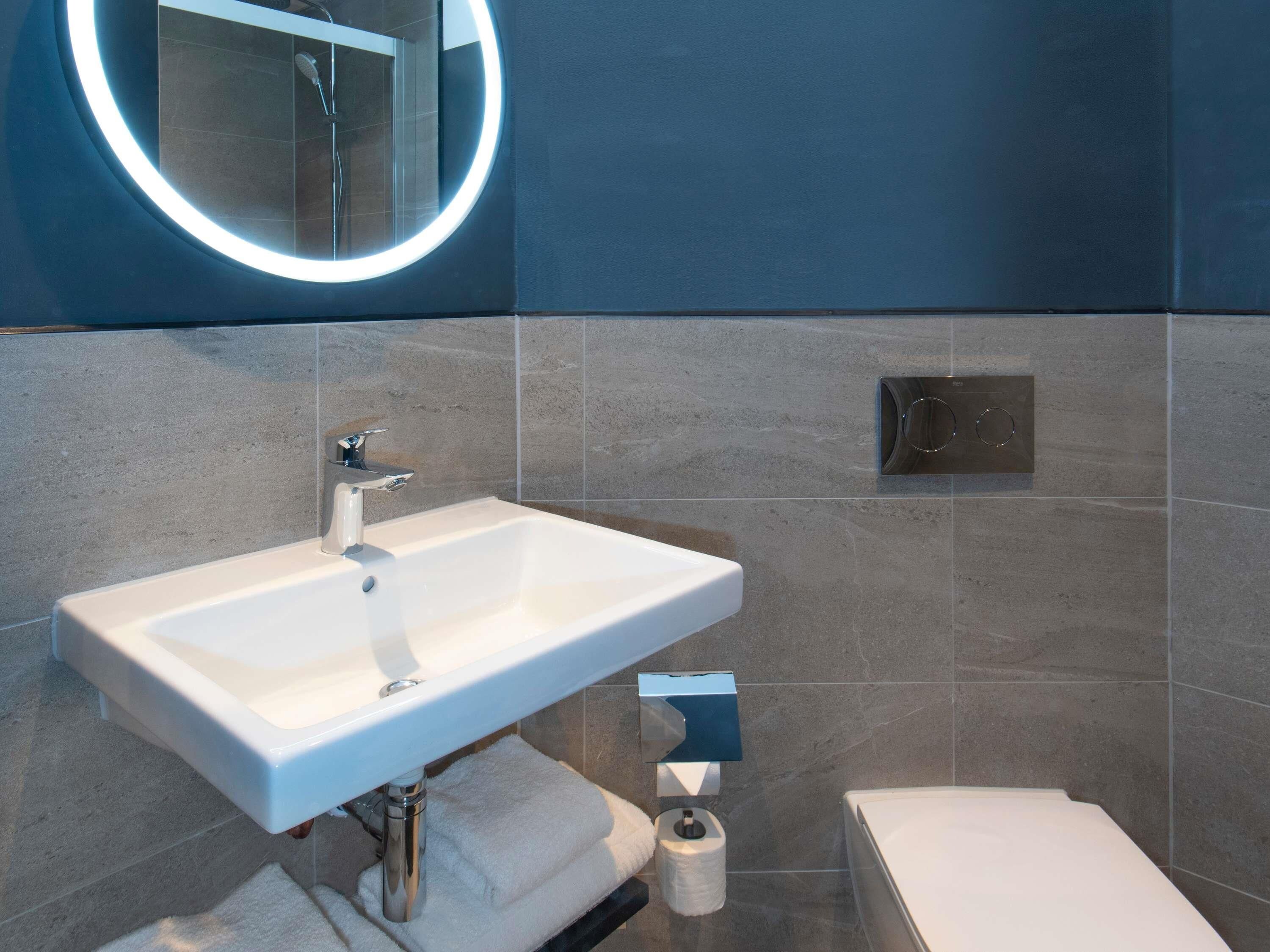 A modern bathroom with a white sink, a mirror with a circular light, and a toilet.