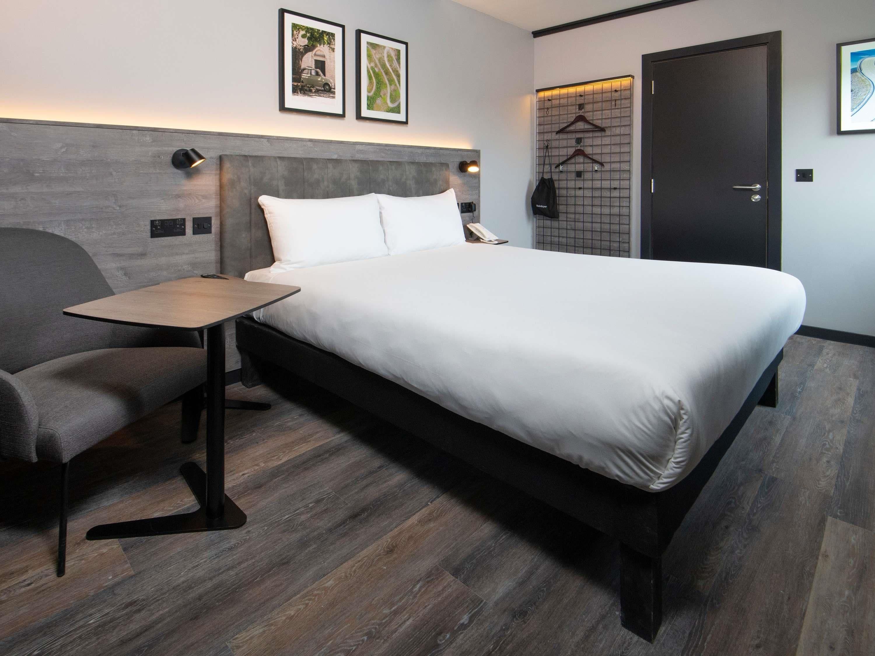 A modern hotel room with a large bed, a small table, a chair, a wardrobe, and a door.