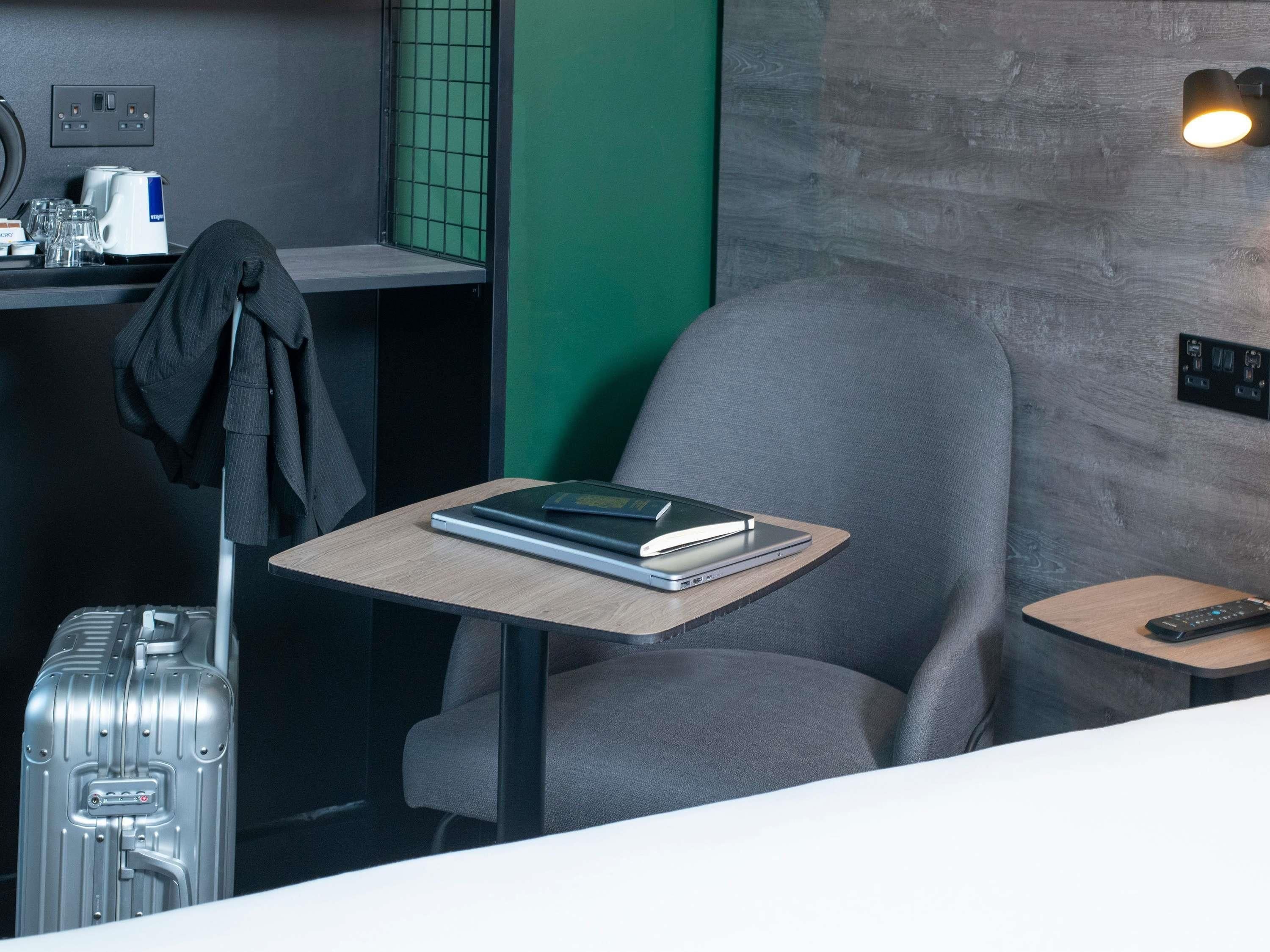 A modern hotel room with a grey armchair, a small wooden table with a book and a remote control, a suitcase, and a coffee machine on a counter.