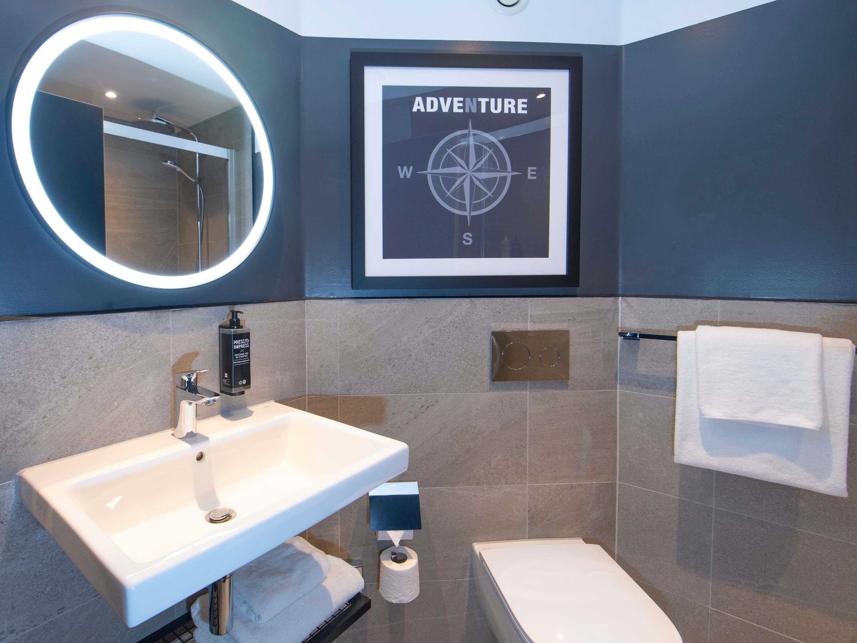 A bathroom with a white sink, a mirror with a circular light, a framed compass sign, a toilet, and a towel rack.