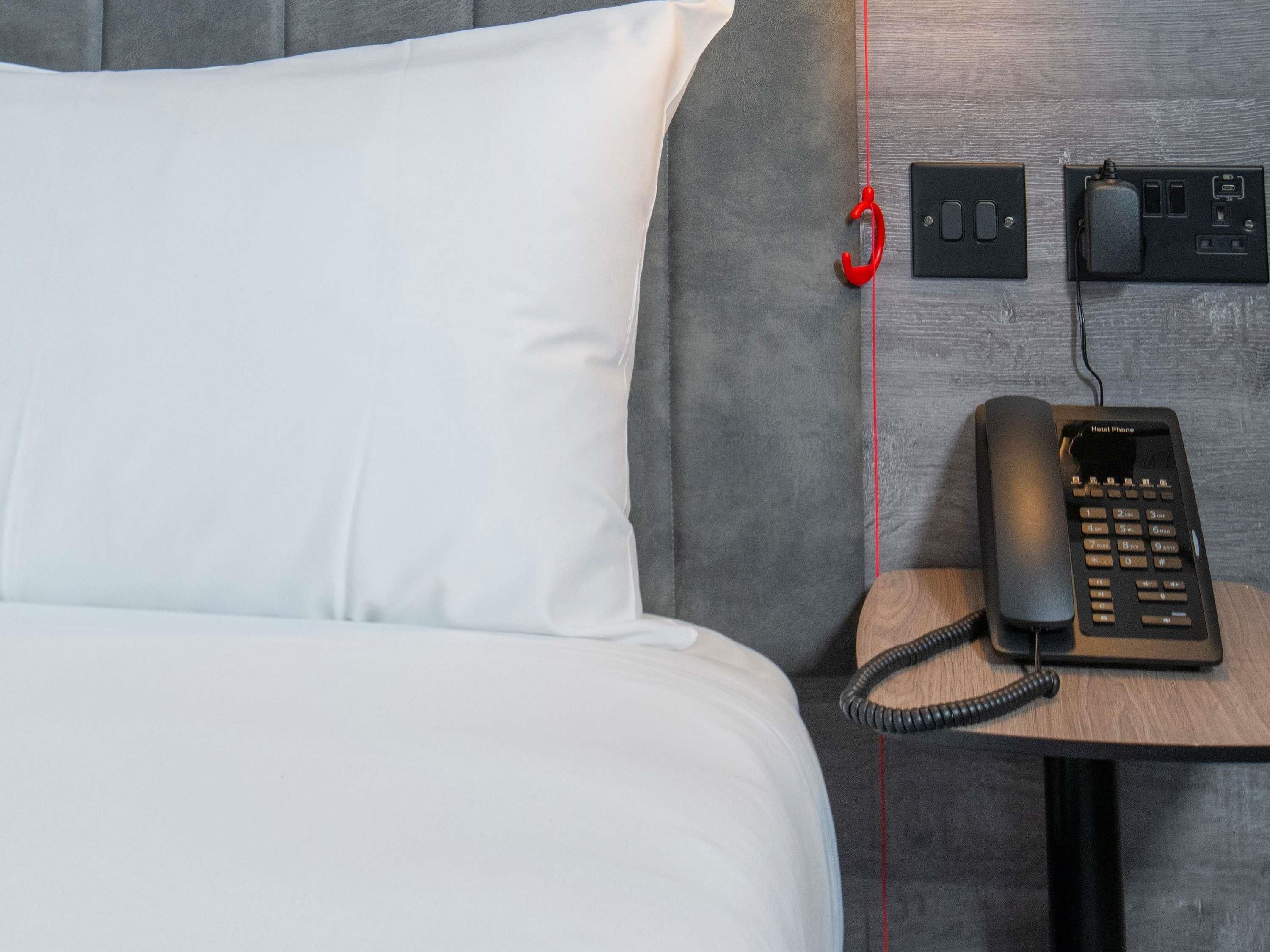 A hotel room with a bed, a bedside table with a telephone, and a power outlet.