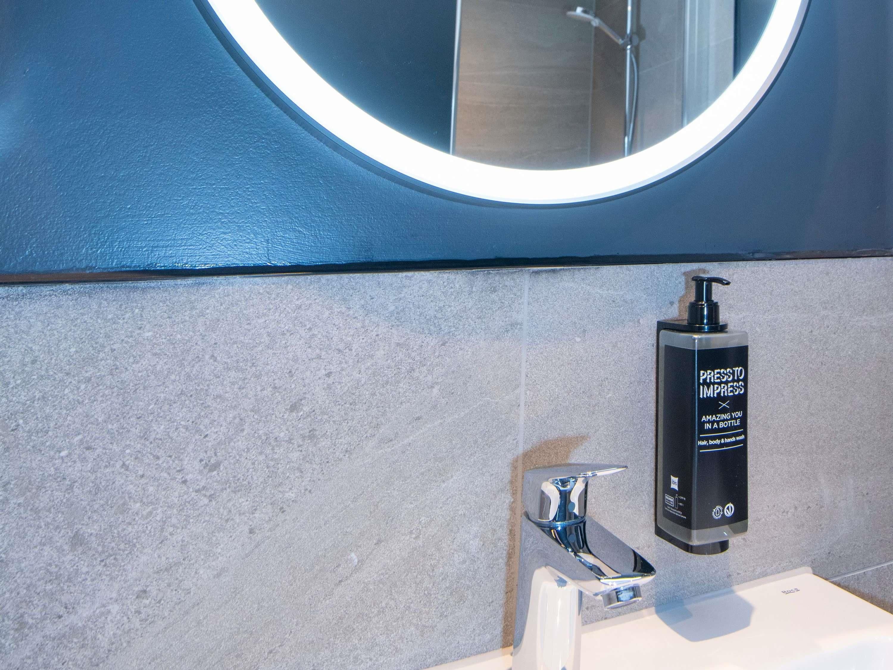 A bathroom with a modern design, featuring a round mirror, a wall-mounted soap dispenser, and a chrome faucet.