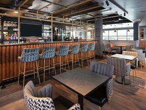 A bar area with a bar counter, multiple stools, and a seating area with tables and chairs. - Ibis Styles London Gatwick Airport (Crawley)