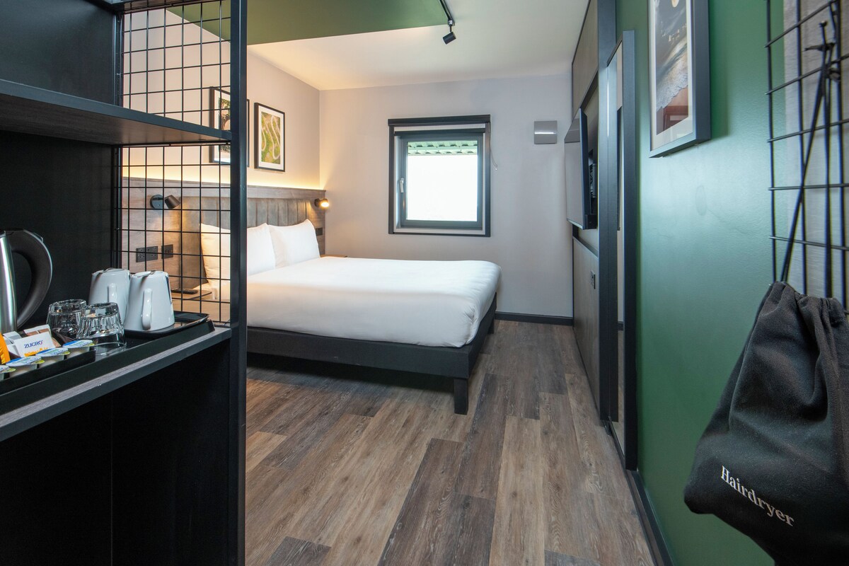 Habitación de hotel compacta con cama, un escritorio pequeño con hervidor, vasos y secador de pelo.