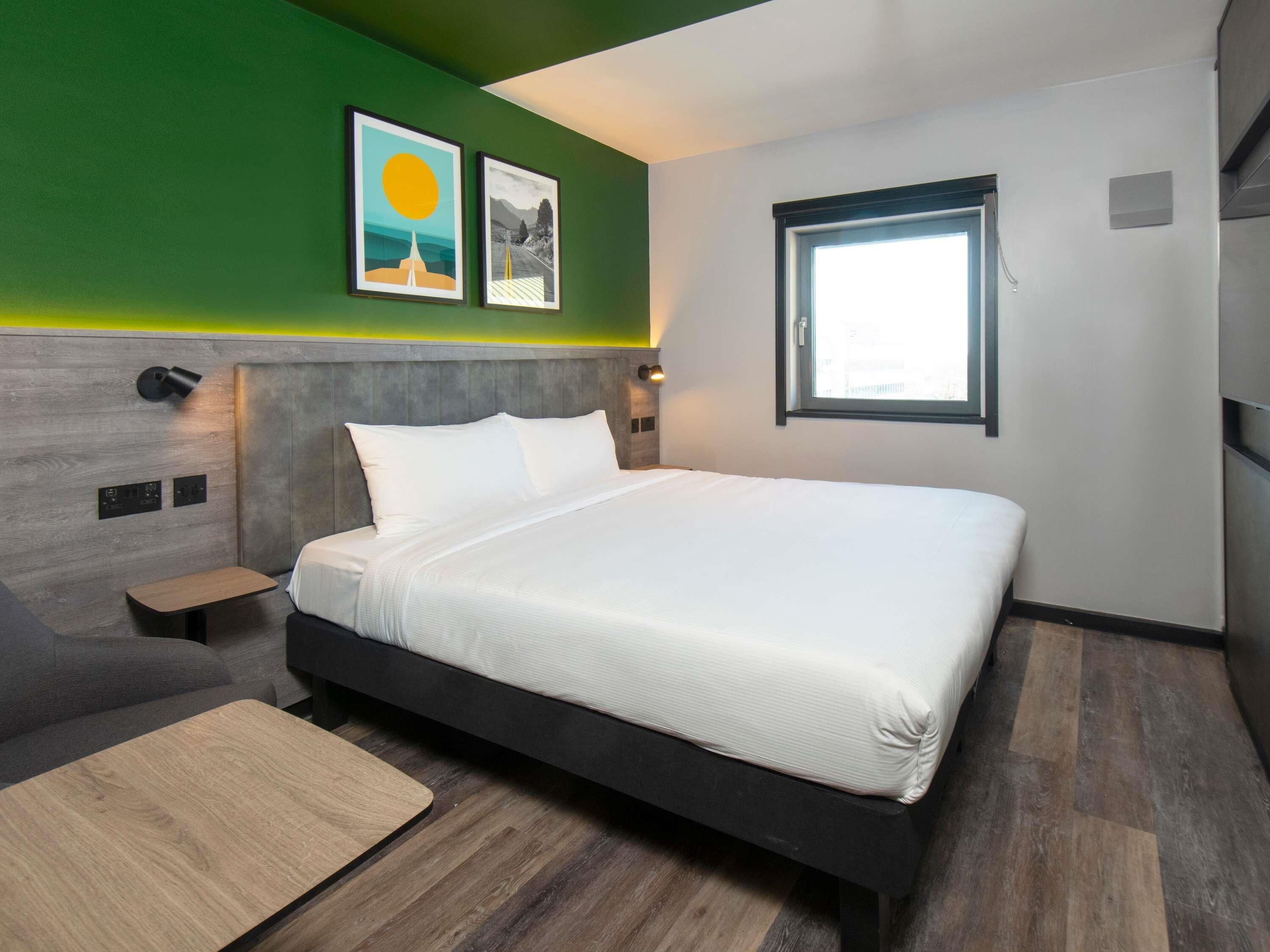 A modern hotel room with a bed, a small table, a nightstand, a window, and artwork on the wall.