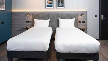 Hotellihuone, jossa on kaksi erillistä sänkyä, harmaa pääty ja kaksi kehystettyä kuvaa seinällä.